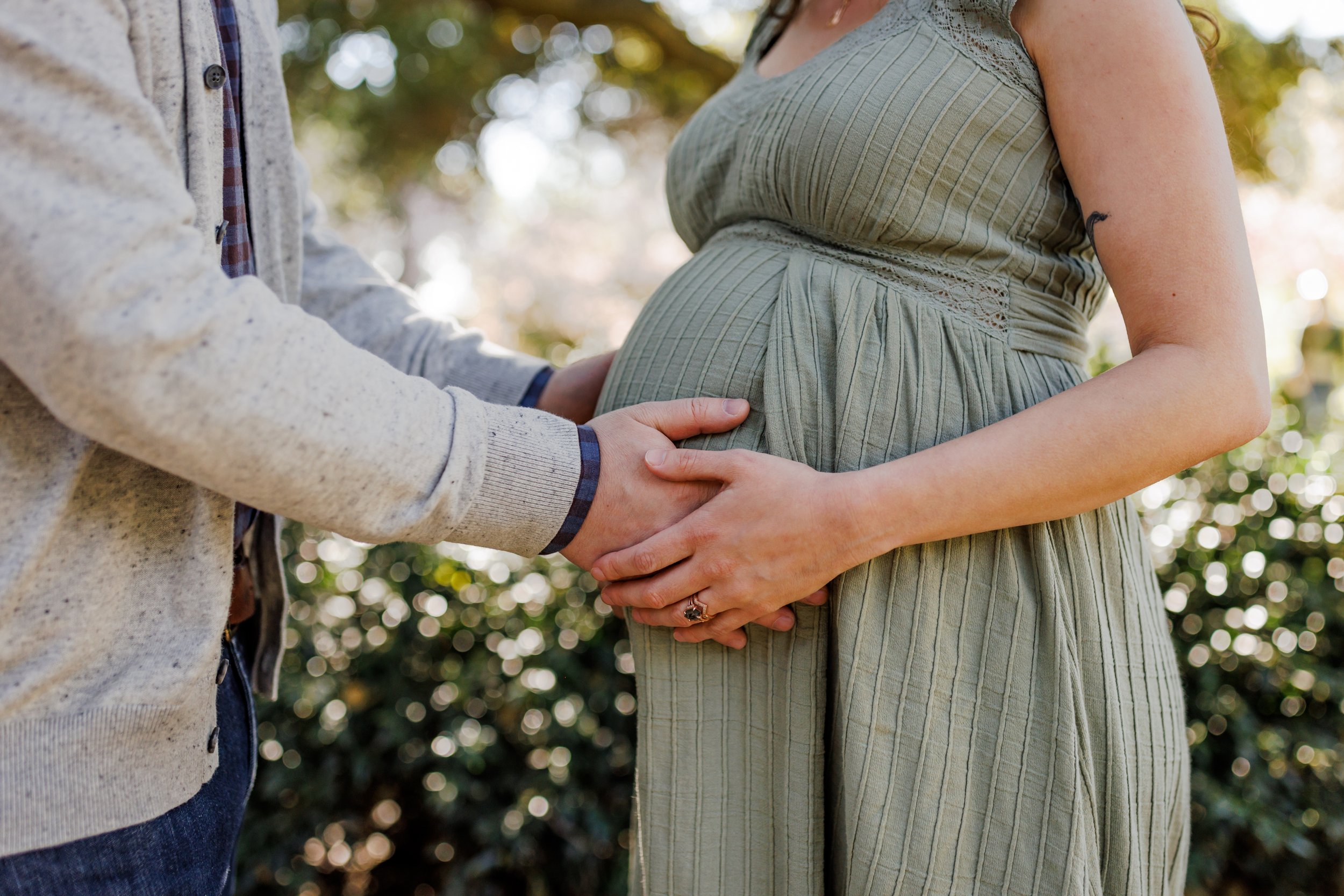 pregnant woman in green dress facing husband with both of their hands on baby bump in garden