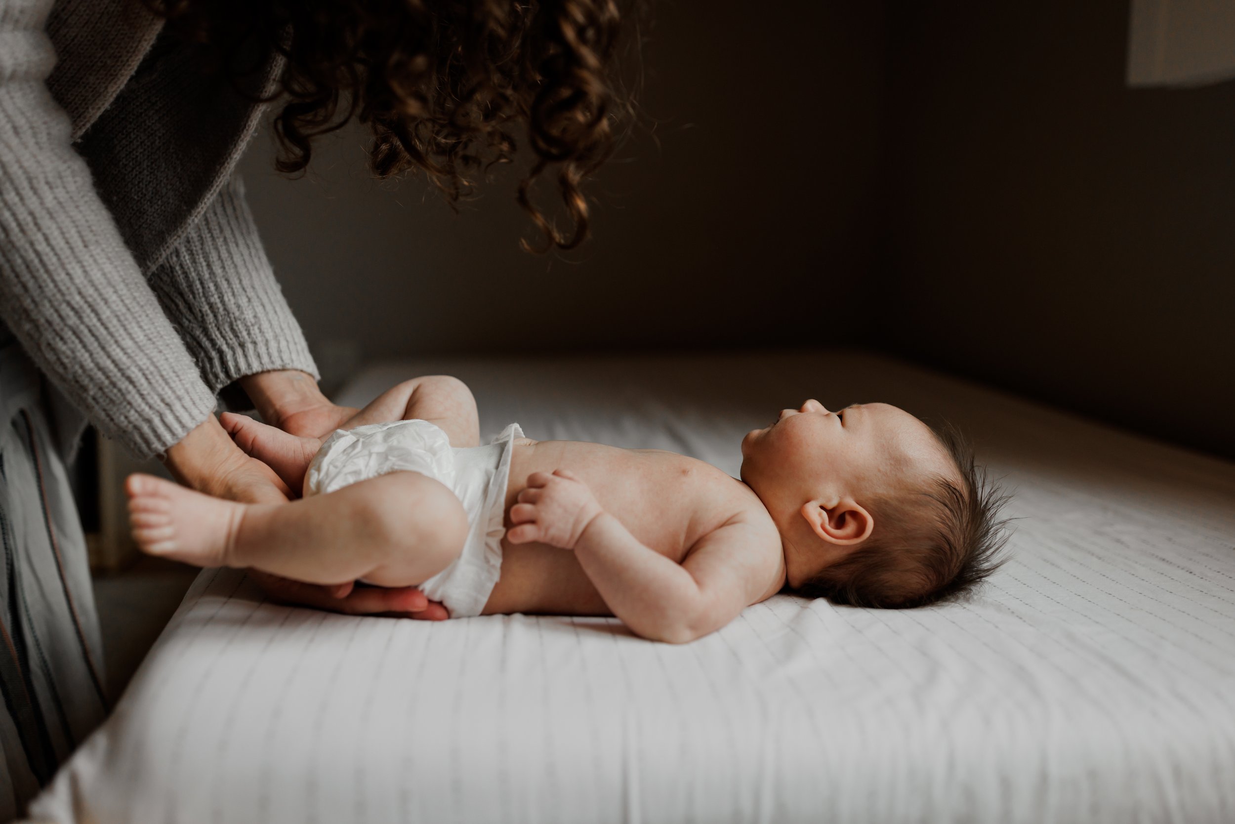 newborn laying on bed at home looking up at mom who is leaning over her and changing her diaper