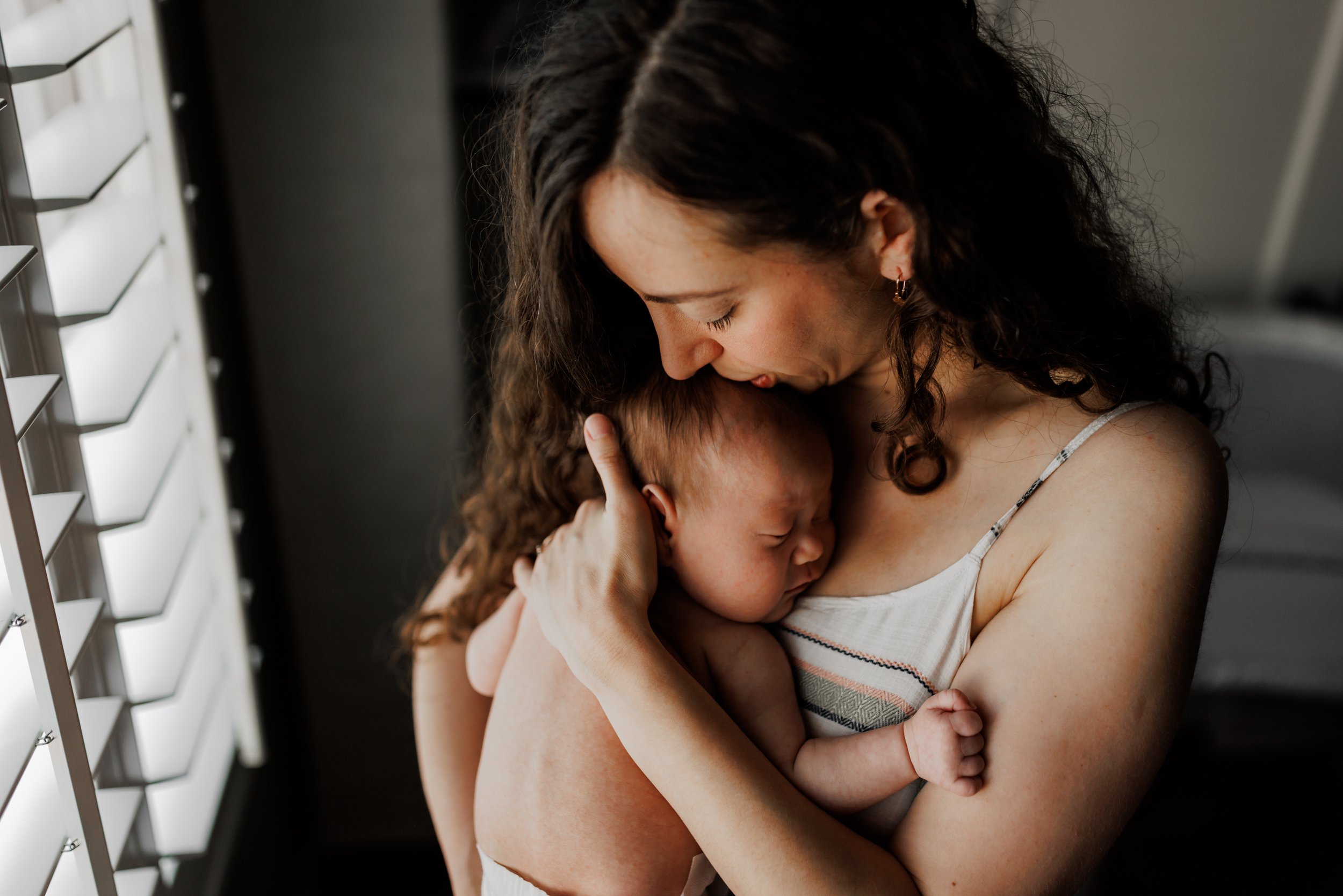 mom holding baby close and kissing her head while standing next to window