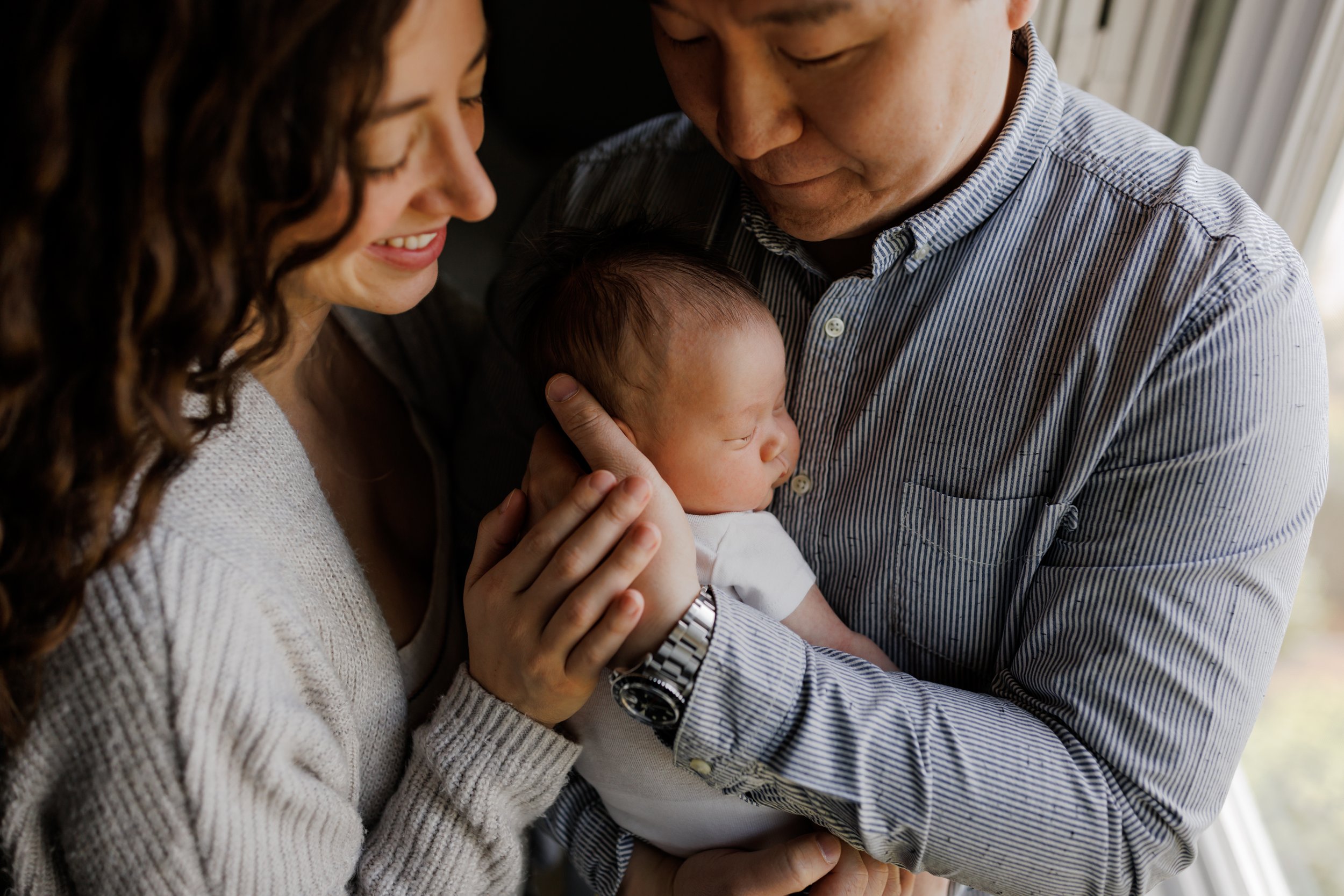 mom and dad standing close touching hands to hold newborn