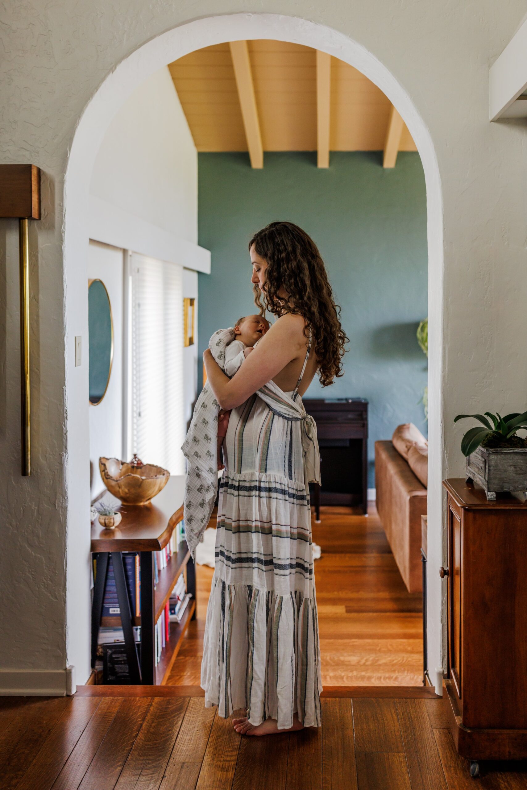 mom standing in arched doorway in striped dress holding newborn at home