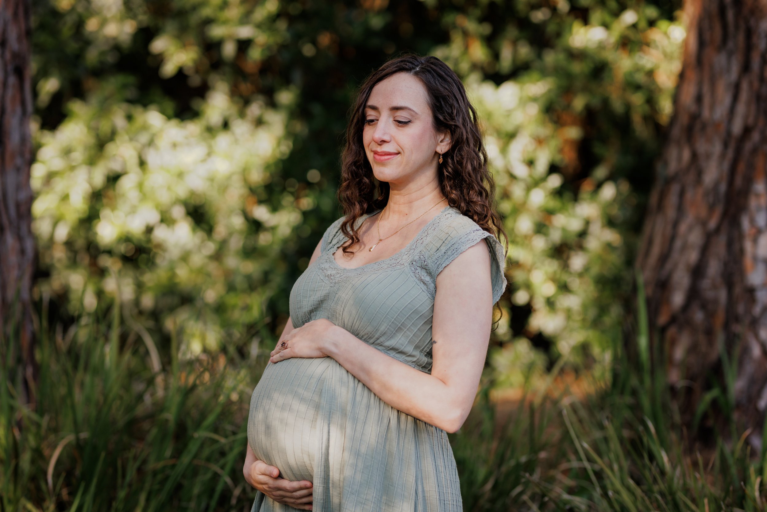 pregnant woman in green dress standing in woods with head down slightly and hands cradling her baby bump