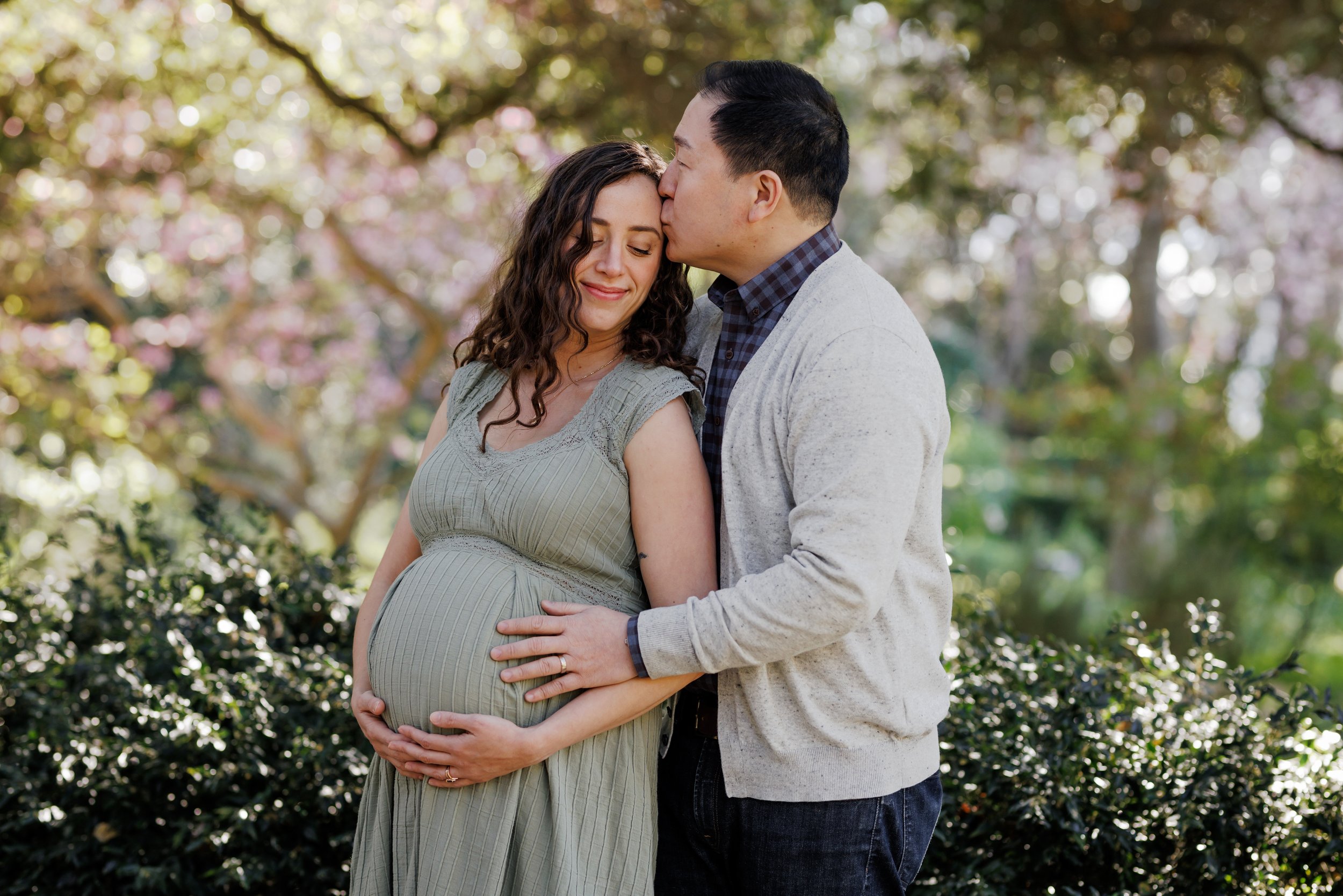 pregnant mom outdoors snuggling up close to her husband resting his hands on her bump standing in front of greenery