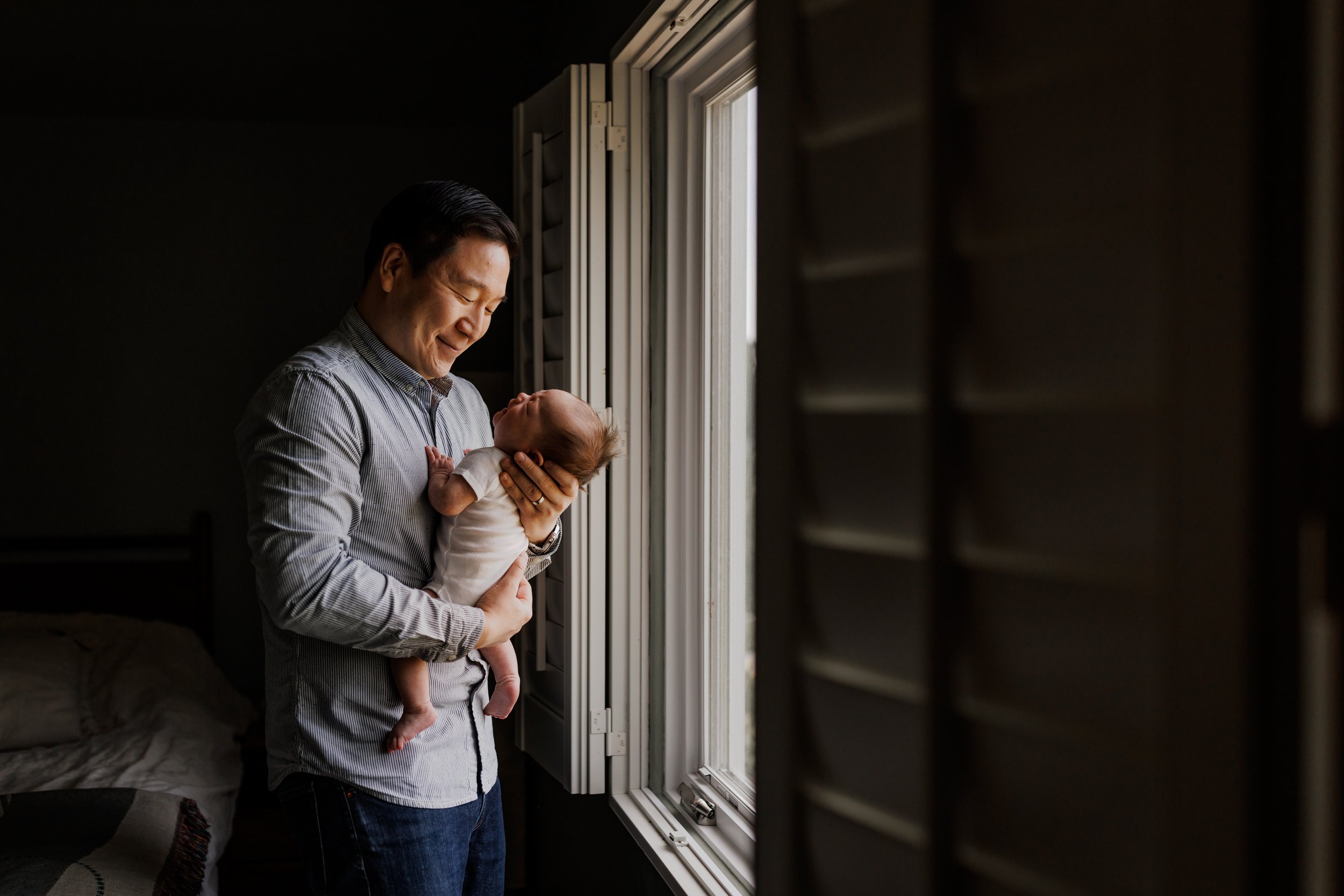 dad looking down and smiling while holding newborn next to window