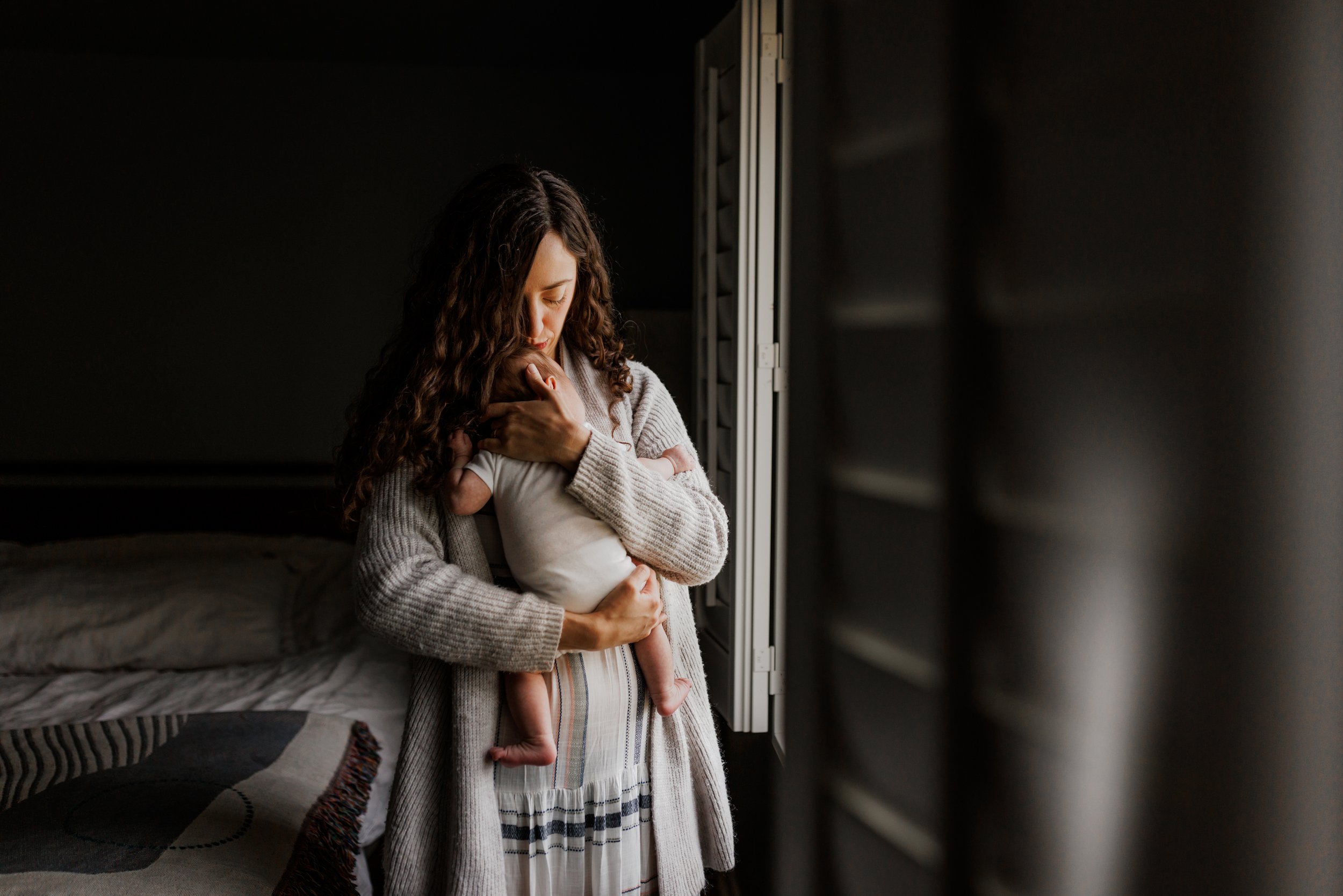 mom holding and kissing newborn baby while standing next to a window