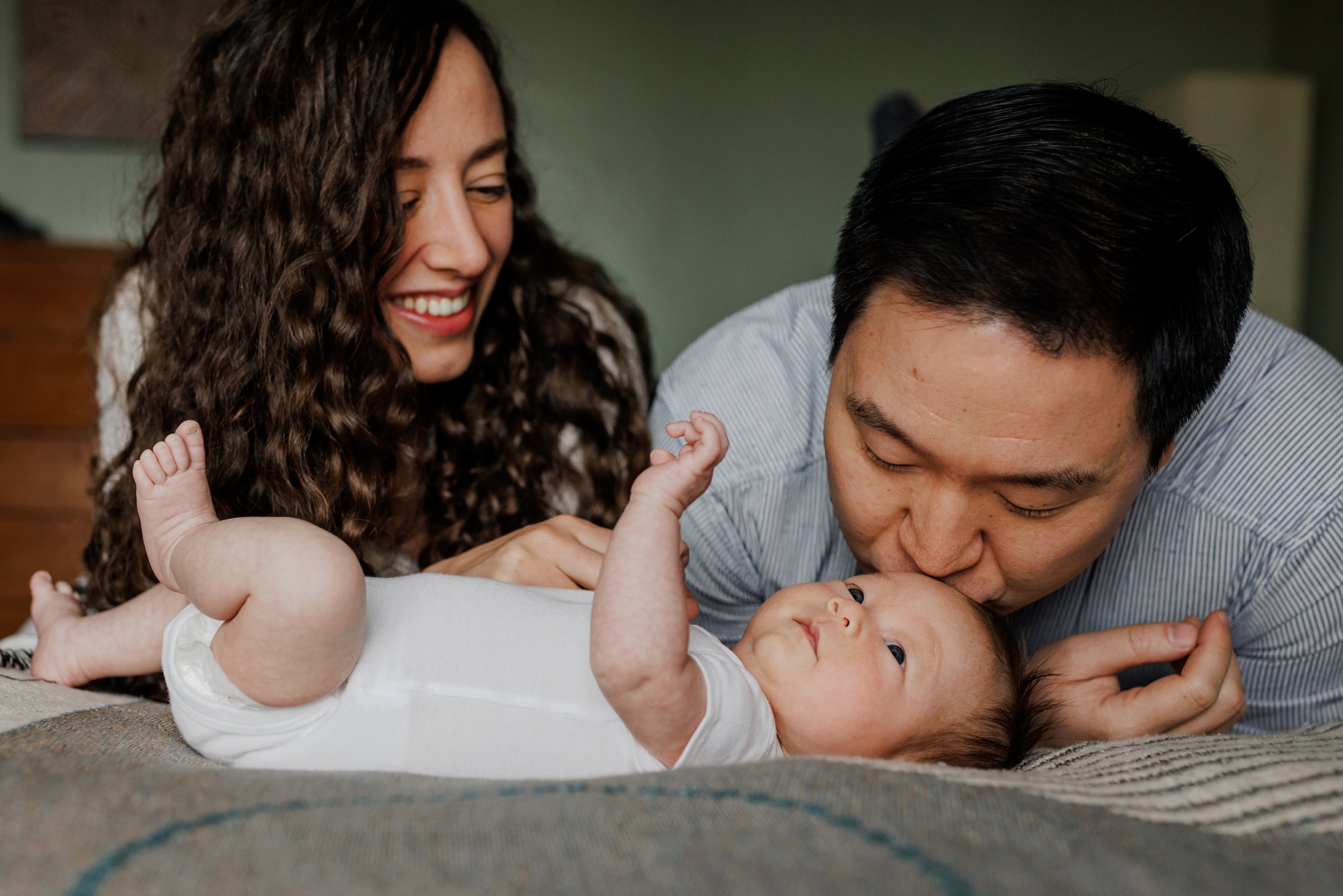 parents leaning over newborn at home on bed as dad kisses baby's head