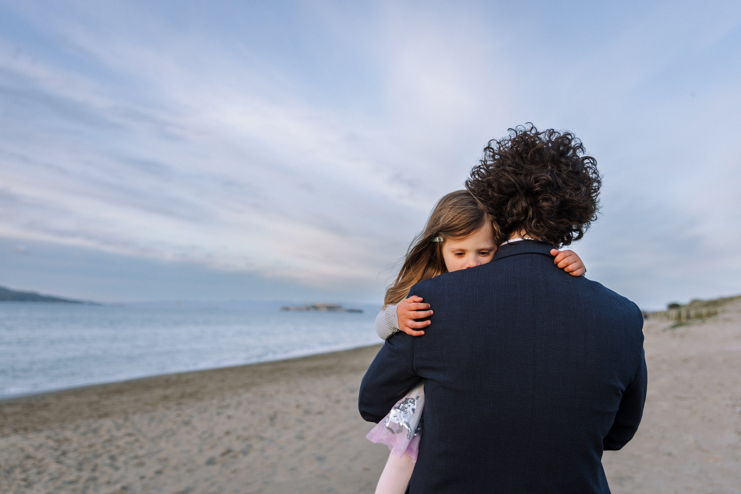 little girl snuggled up on dad's shoulder on the beach as he walks