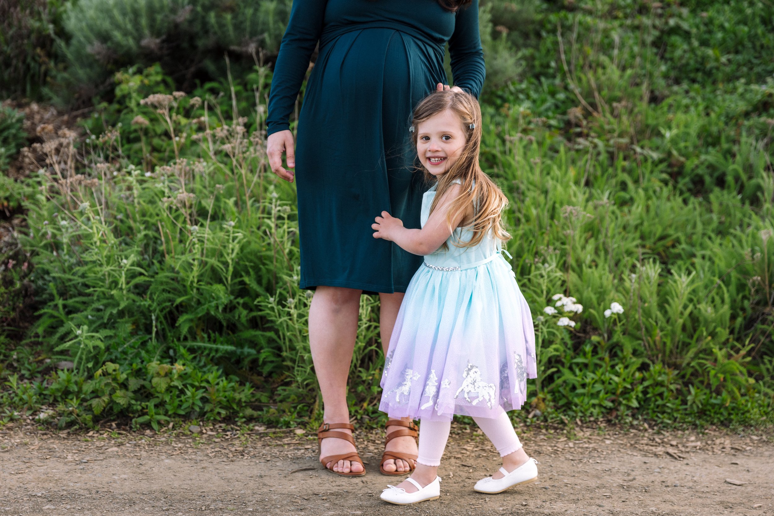 4-year-old girl standing by pregnant mom and looking at the camera and smiling at a park