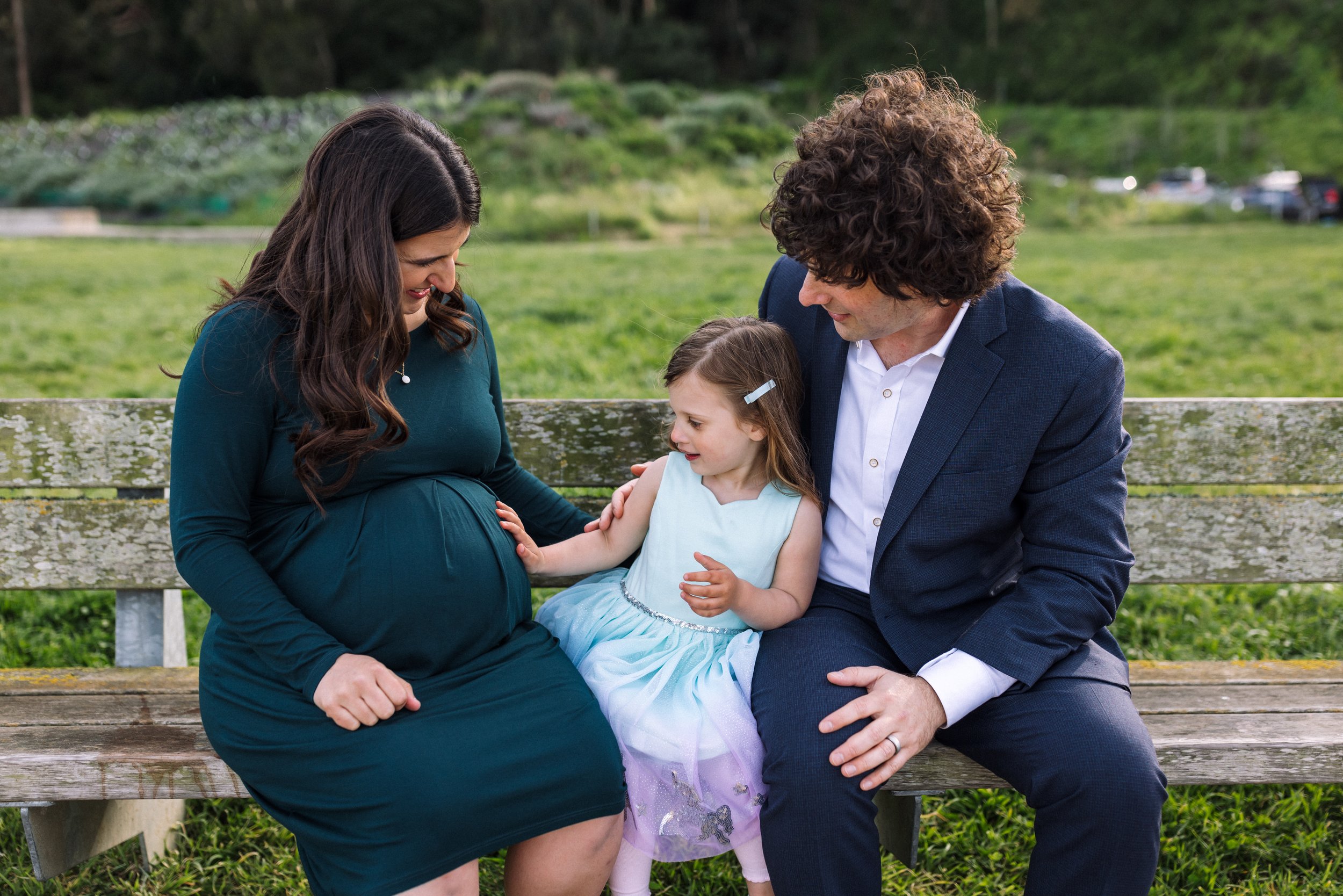 pregnant mom sitting on bench with her daughter and husband and little girl is touching mom's baby bump