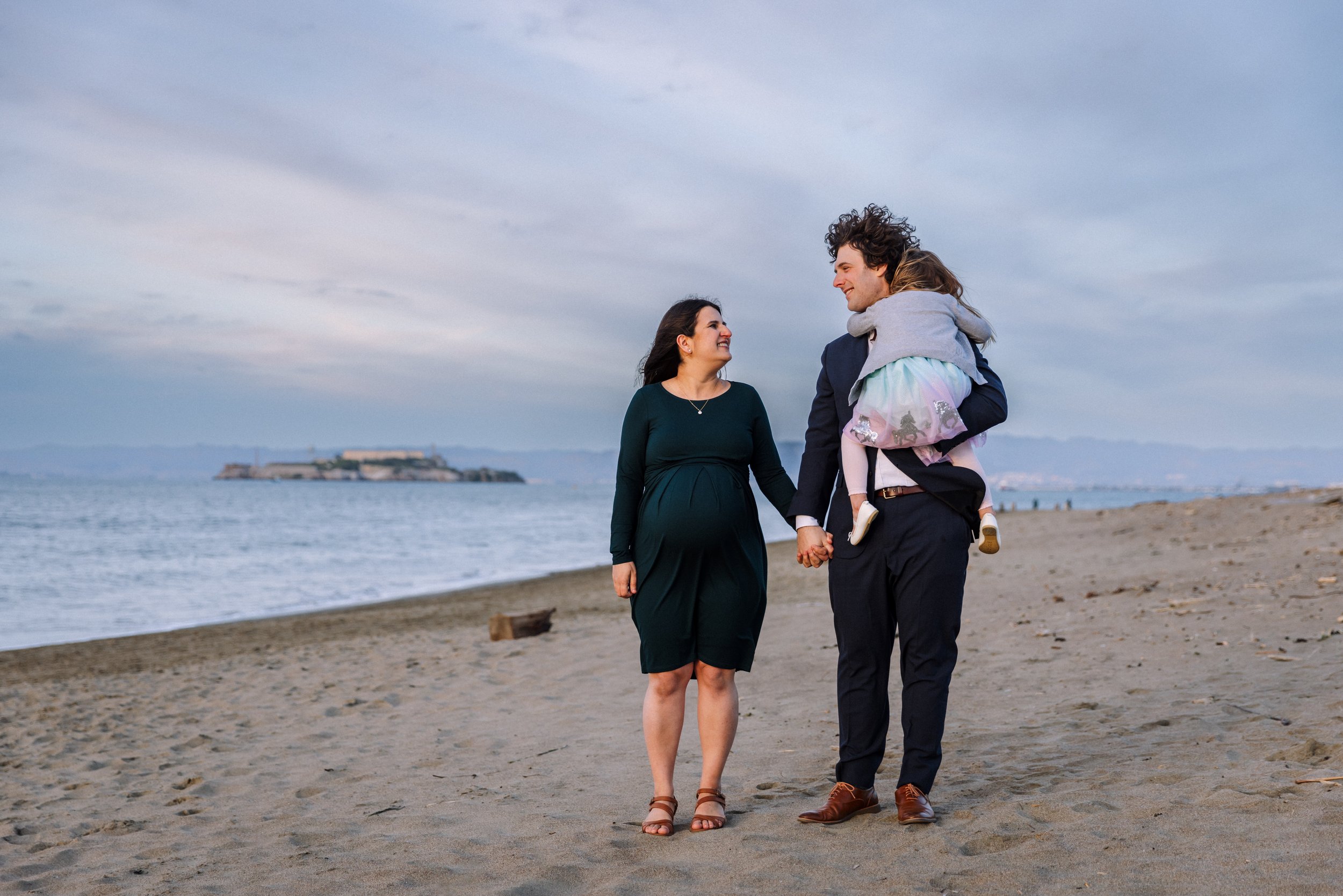 pregnant mom holding hands with husband who is holding daughter and walking together on the beach looking at each other