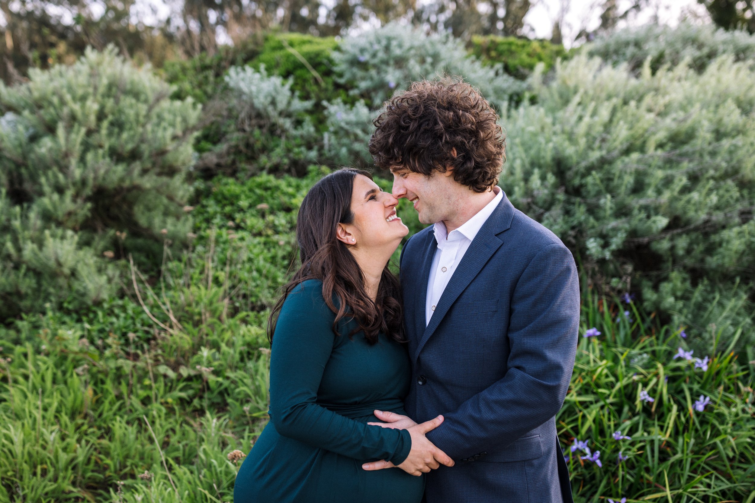 pregnant mom and partner standing nose to nose in front of greenery at a park and smiling at each other