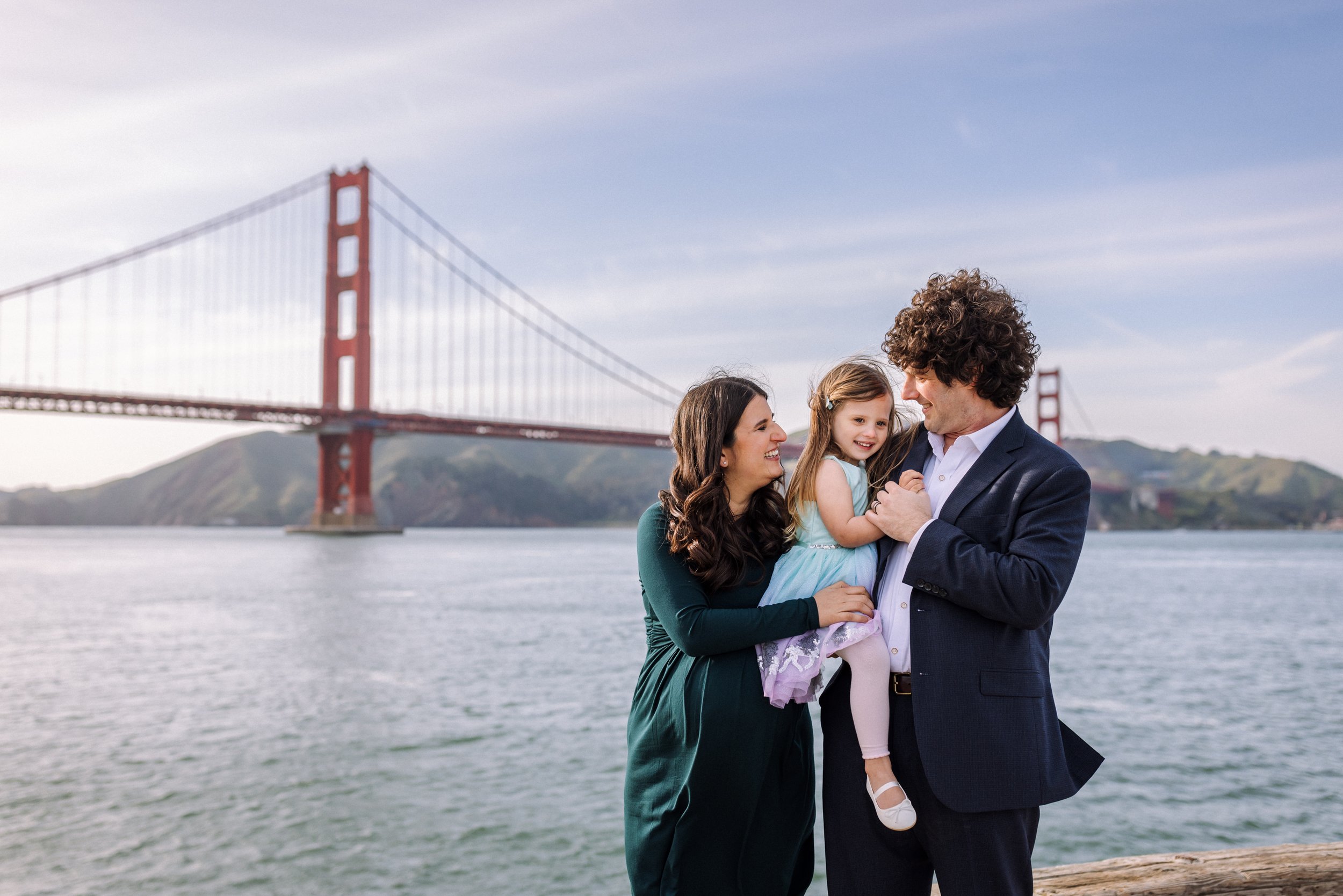 pregnant mom standing and holding 4-year-old daughter with husband in front of golden gate bridge in San Francisco while they both look at her and smile