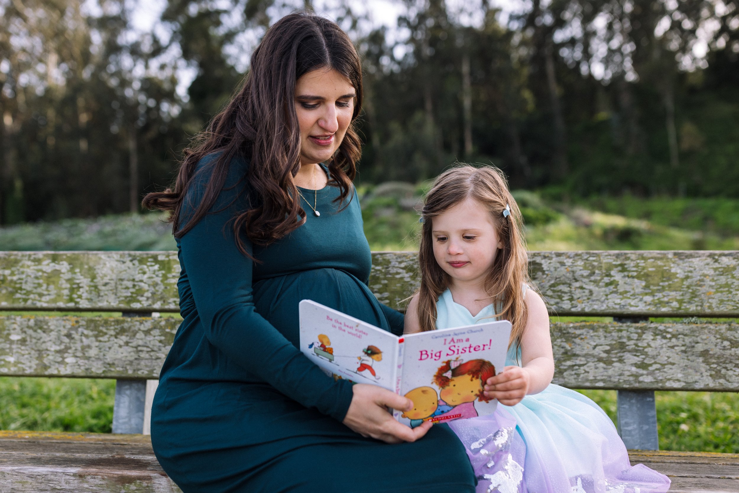 pregnant mom sitting on a bench with four year daughter at a park and reading a book about becoming a big sister