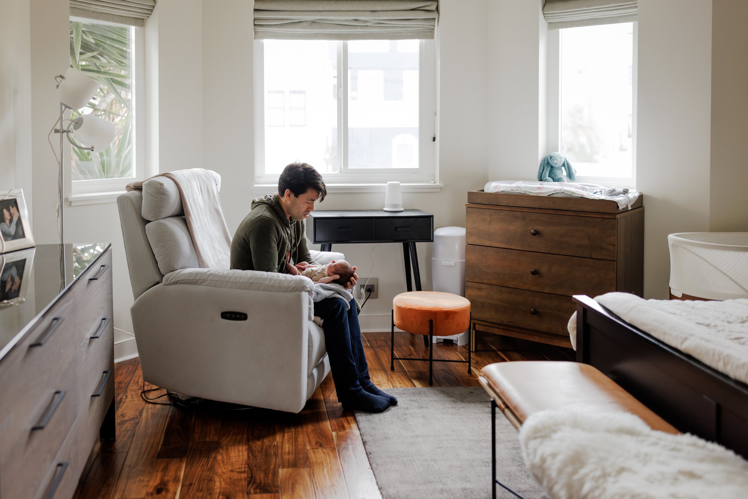 Dad sitting in chair in bedroom holding newborn baby in his lap and looking down at him