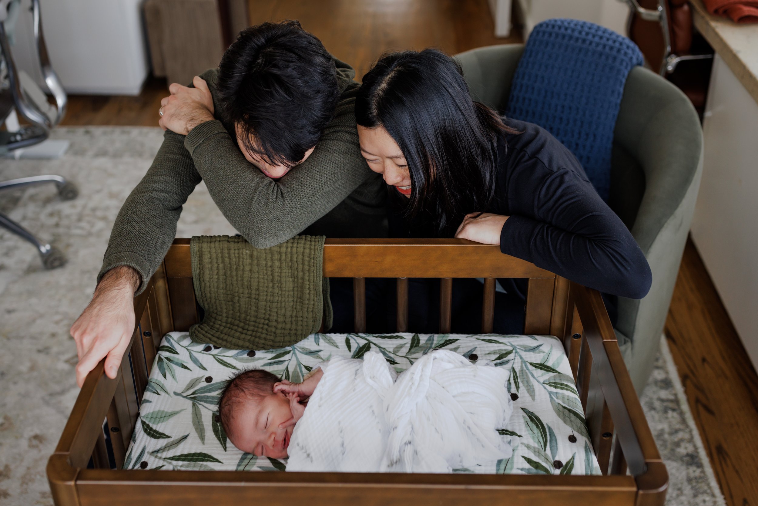 mom and dad leaning over and smiling down at swaddled newborn resting in small crib