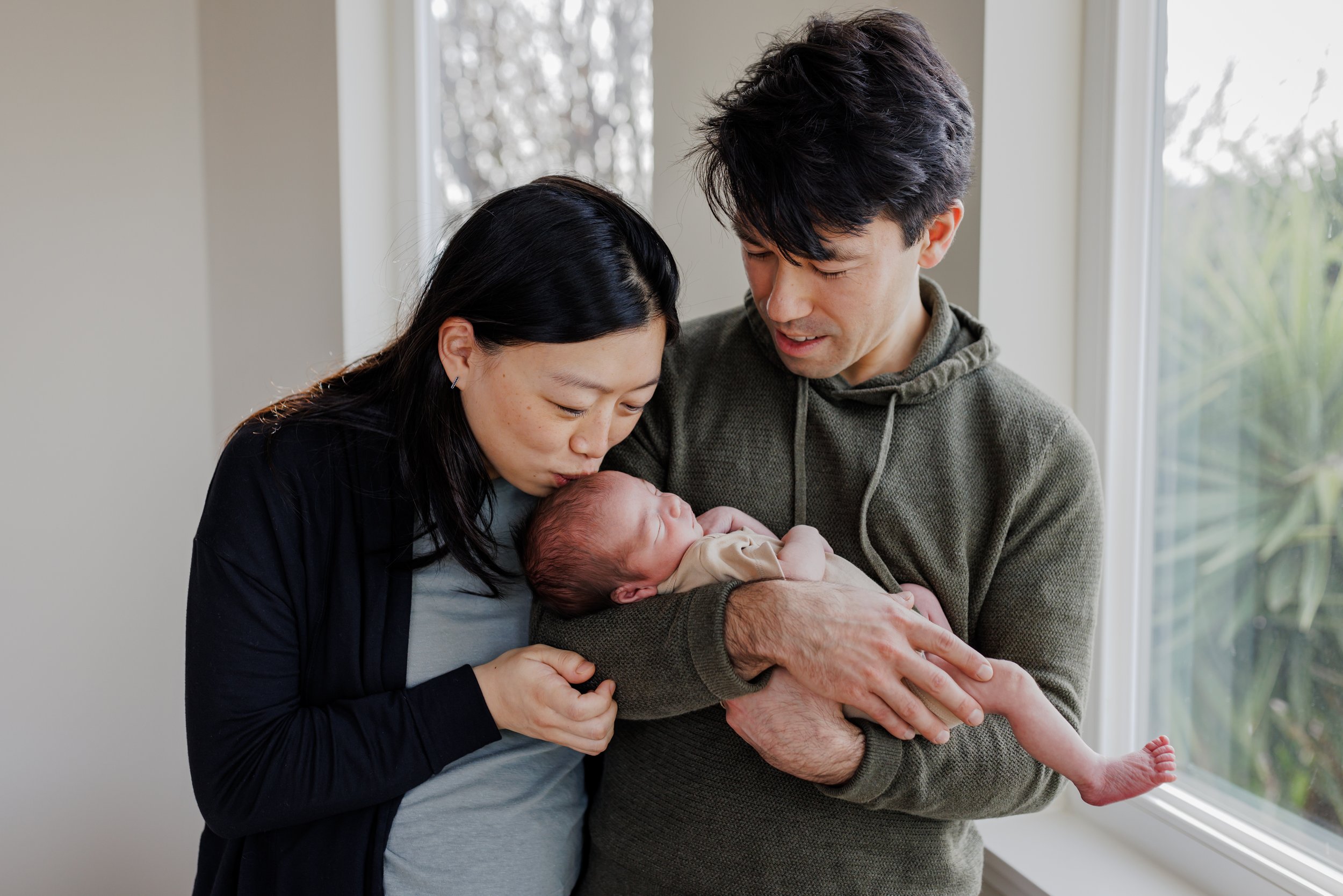 Dad holding newborn in his arms by window and mom is leaning over kissing baby's head