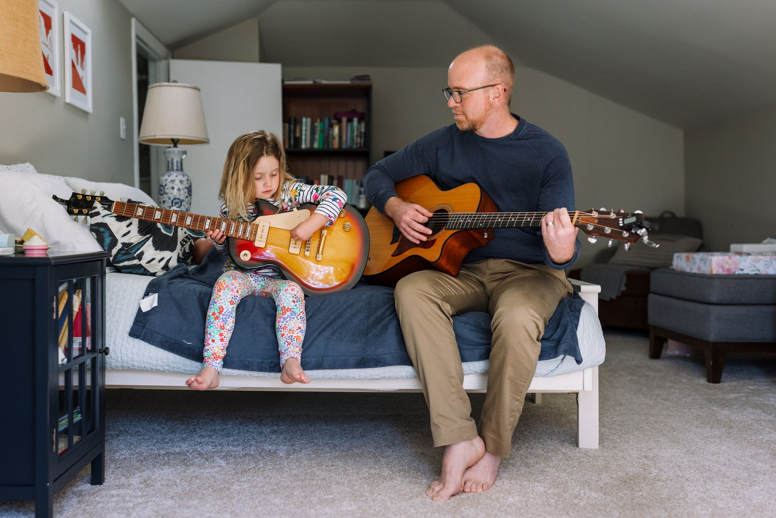 dad-sitting-on-bed-teaching-preschool-daughter-how-to-play-guitar