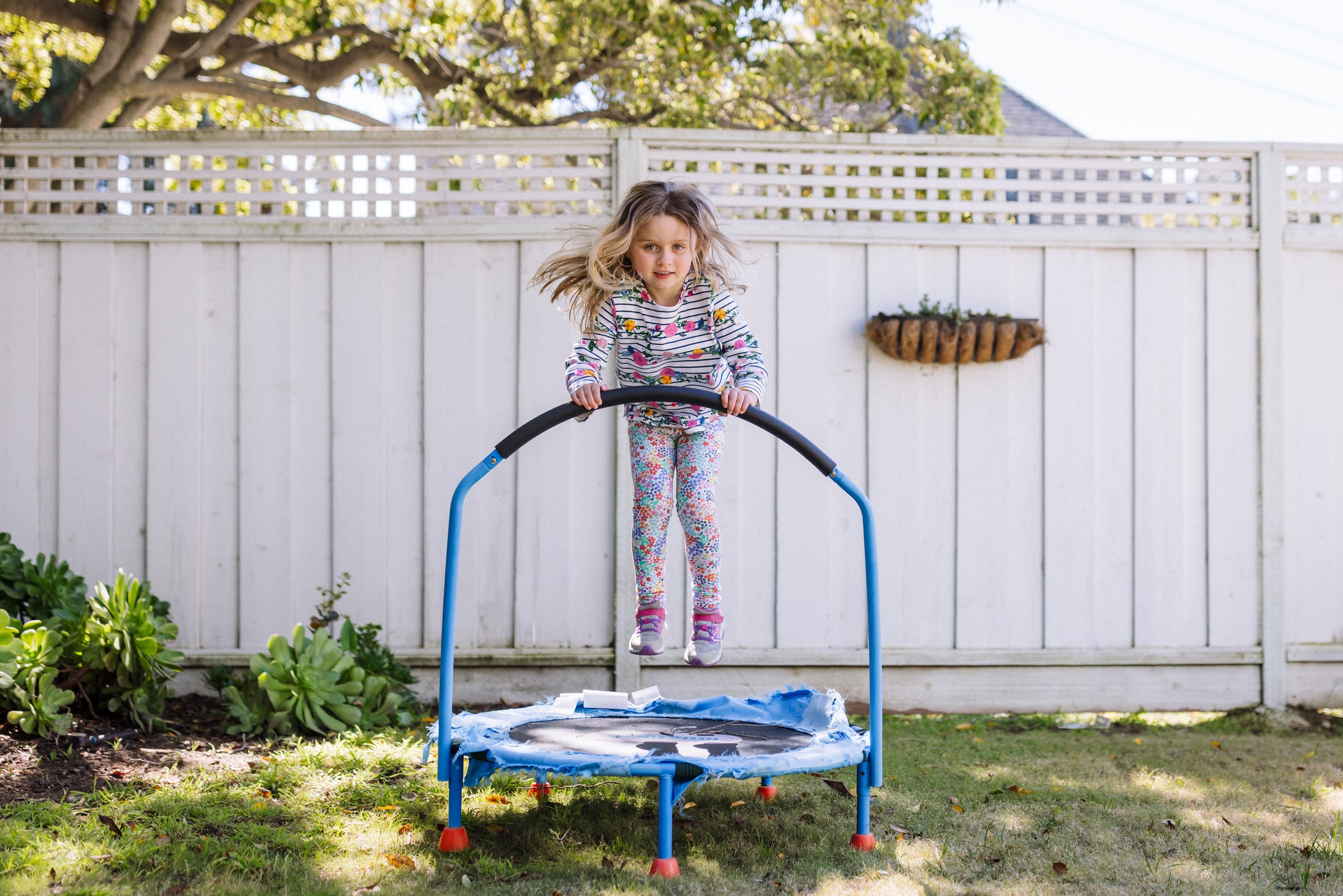 4-year-old girl looking at the camera and smiling as she jumps on small trampoline in the backyard dressed in mismatched clothing