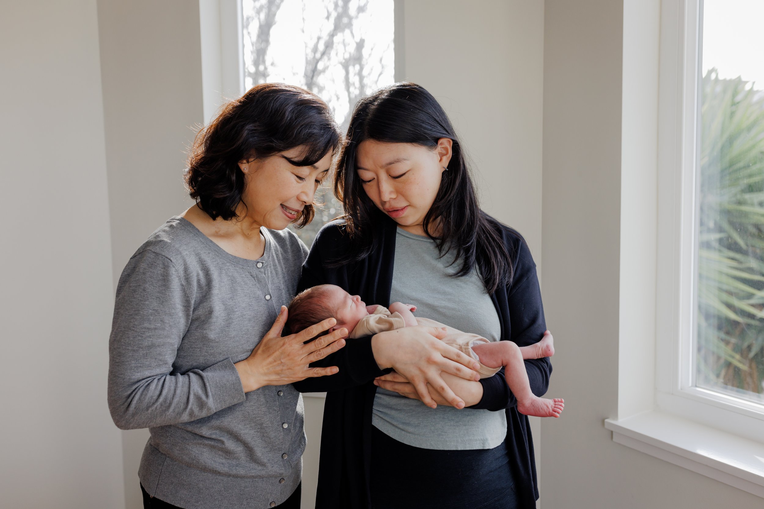 mom holding newborn son as grandma looks down smiling next to her and touching baby's head gently