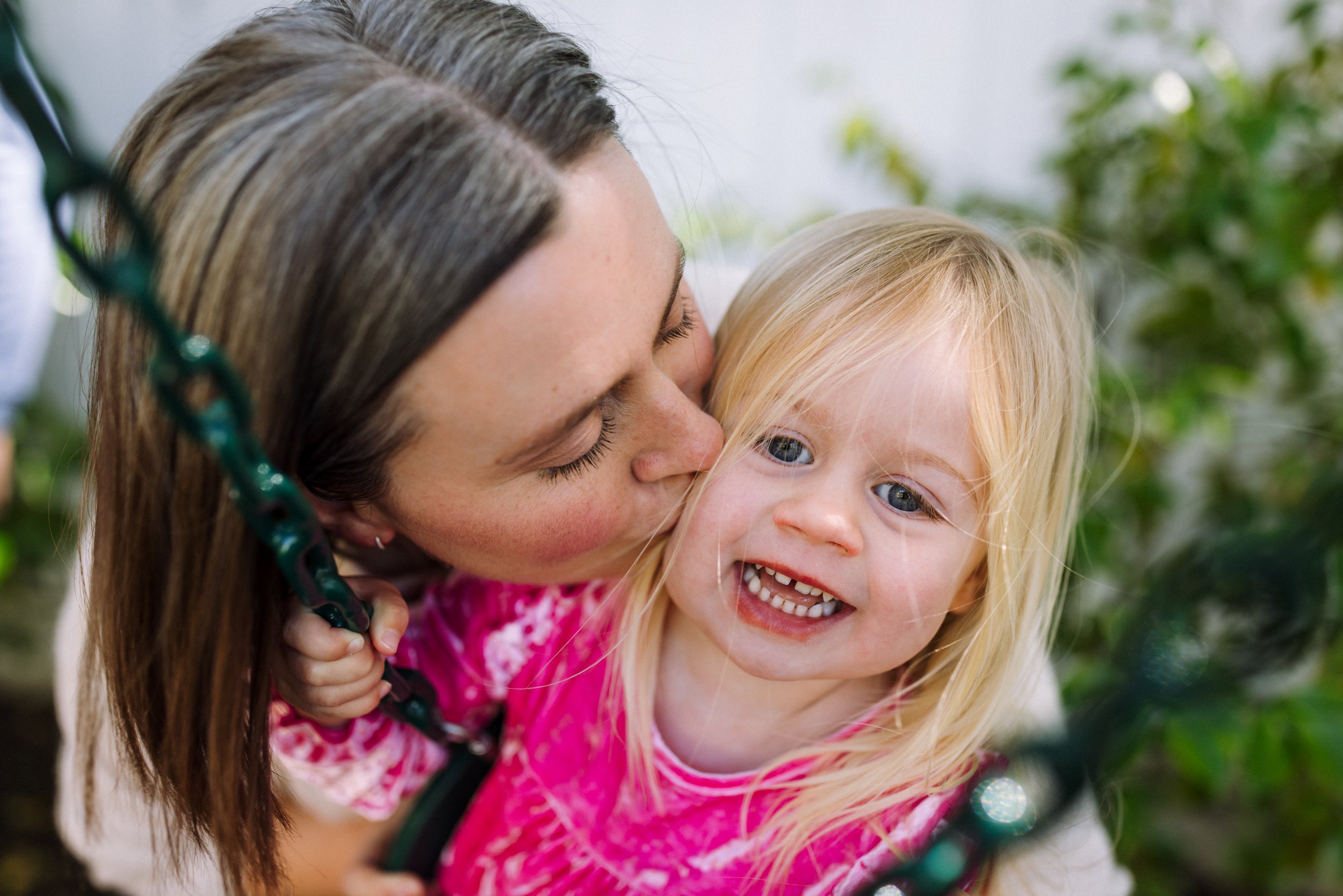 close up of mom kissing toddler daughter while she pauses on the swing