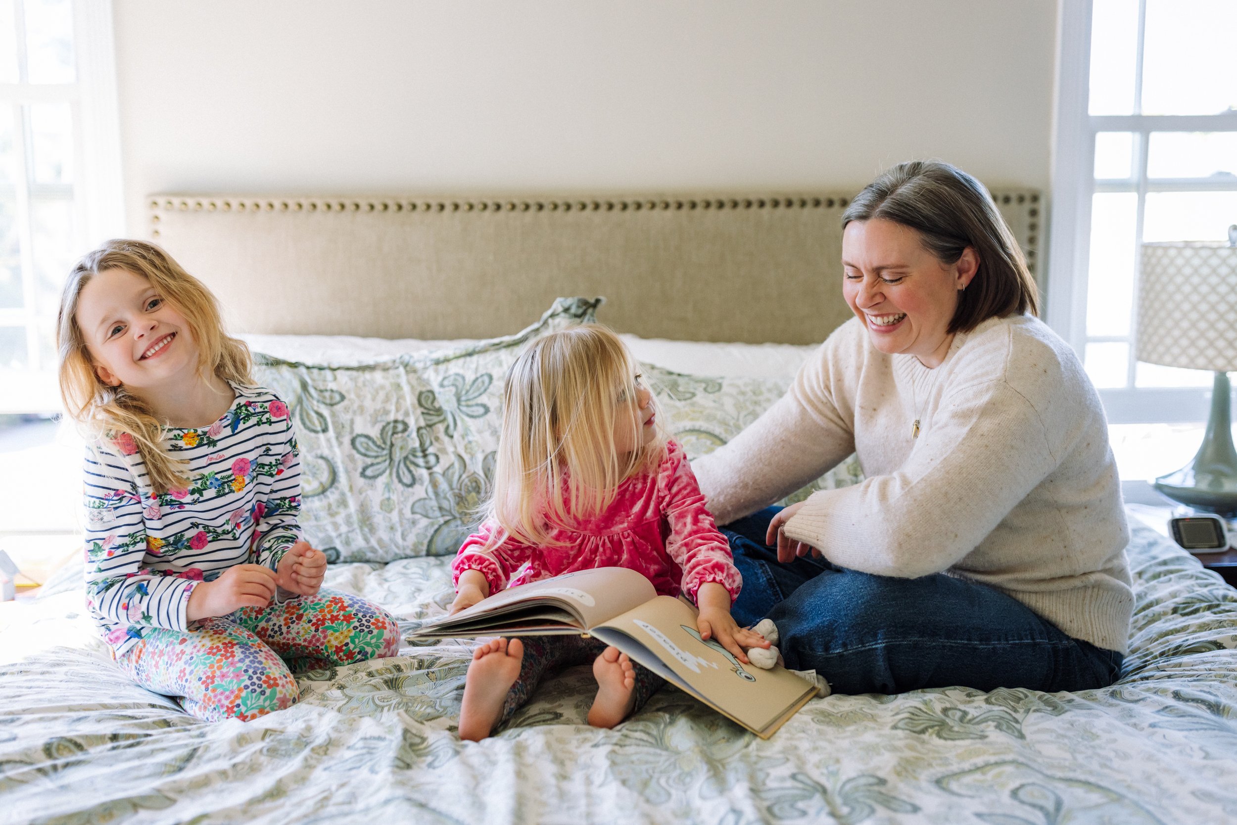 mom sitting on her bed with toddler and preschool daughters laughing together while reading a book