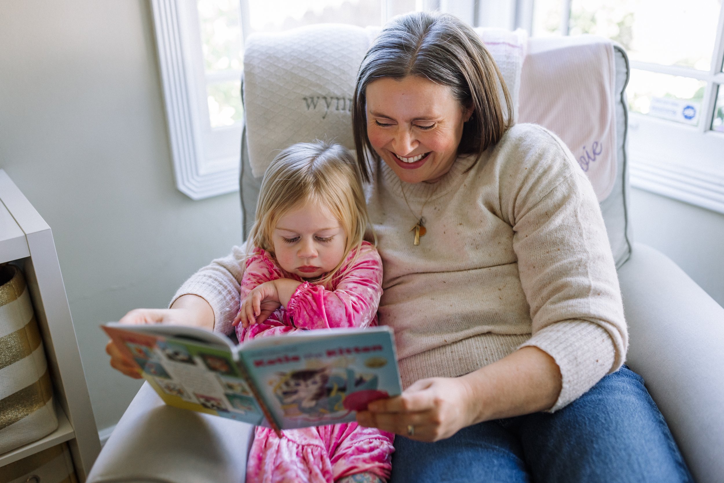 mom sitting in chair in kid's room reading book to toddler daughter during in-home session