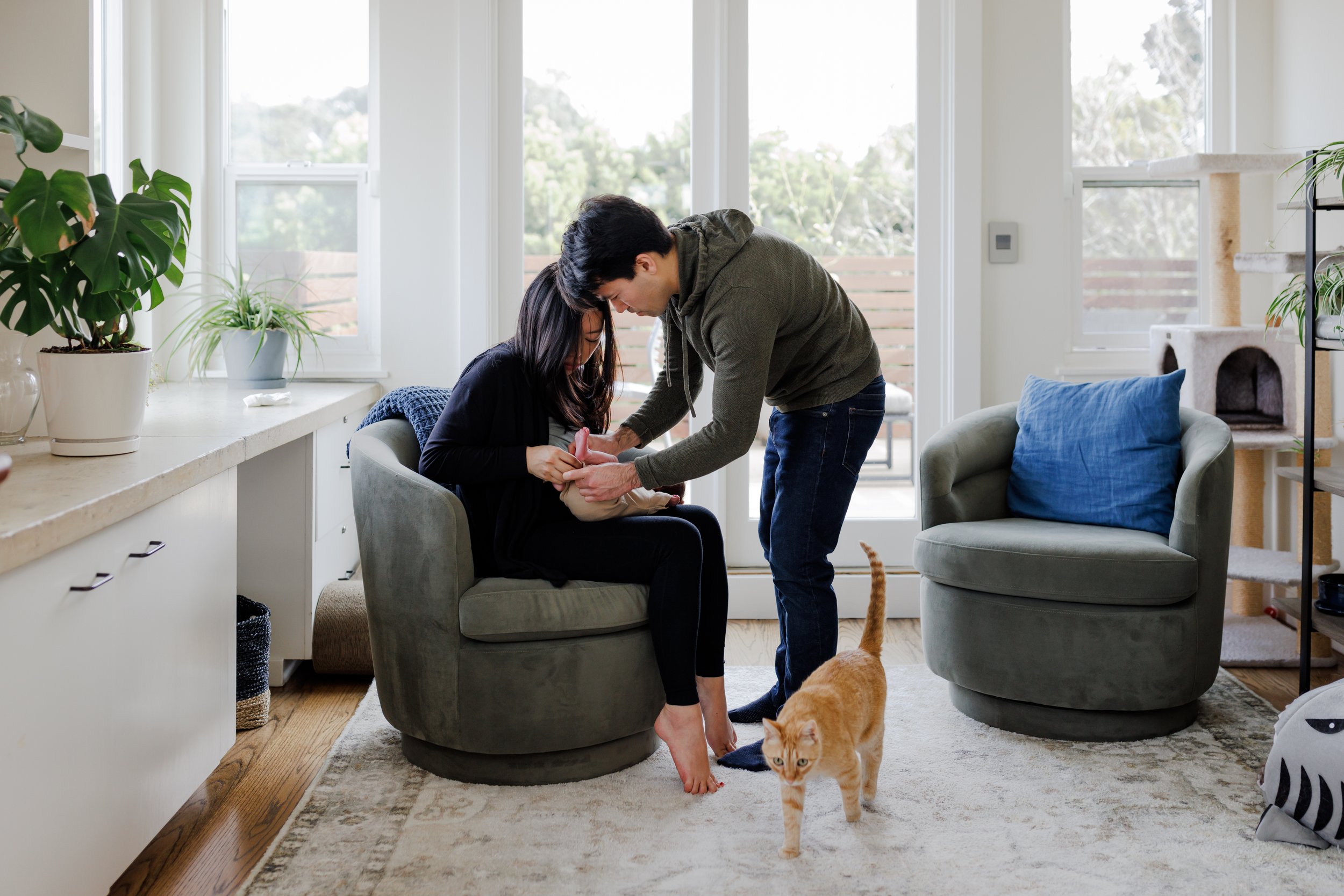 mom sitting in living room chair holding baby while dad stands over helping to change clothes and cat walks in the foreground