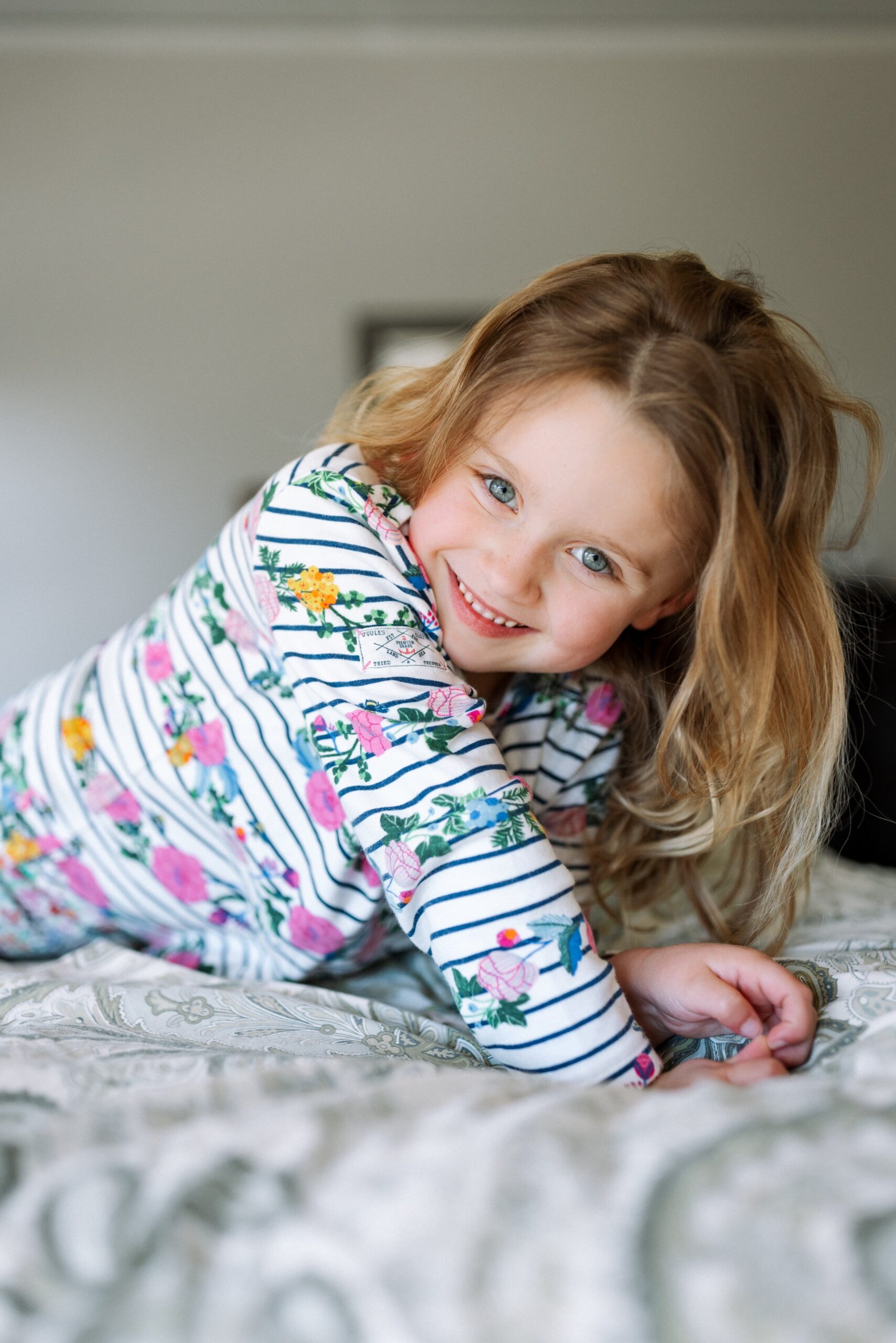 4-year-old girl propping herself up on her parents' bed looking at the camera with big smile dressed in a striped and flower shirt