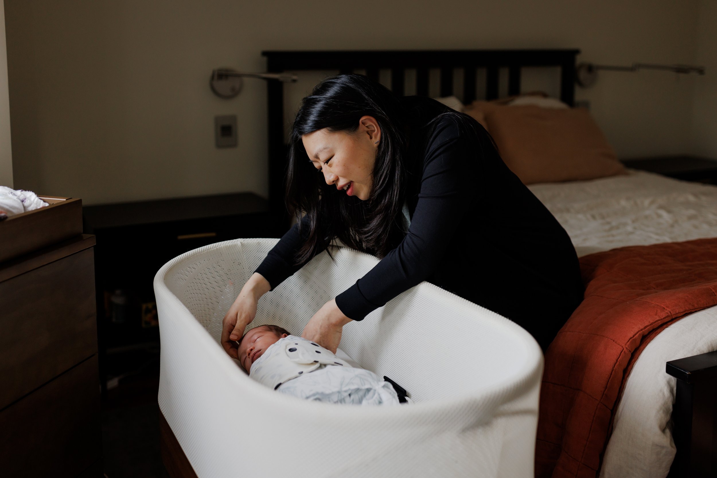 mom leaning over bassinet while sitting on edge of her bed to check on her sleeping and swaddled newborn baby