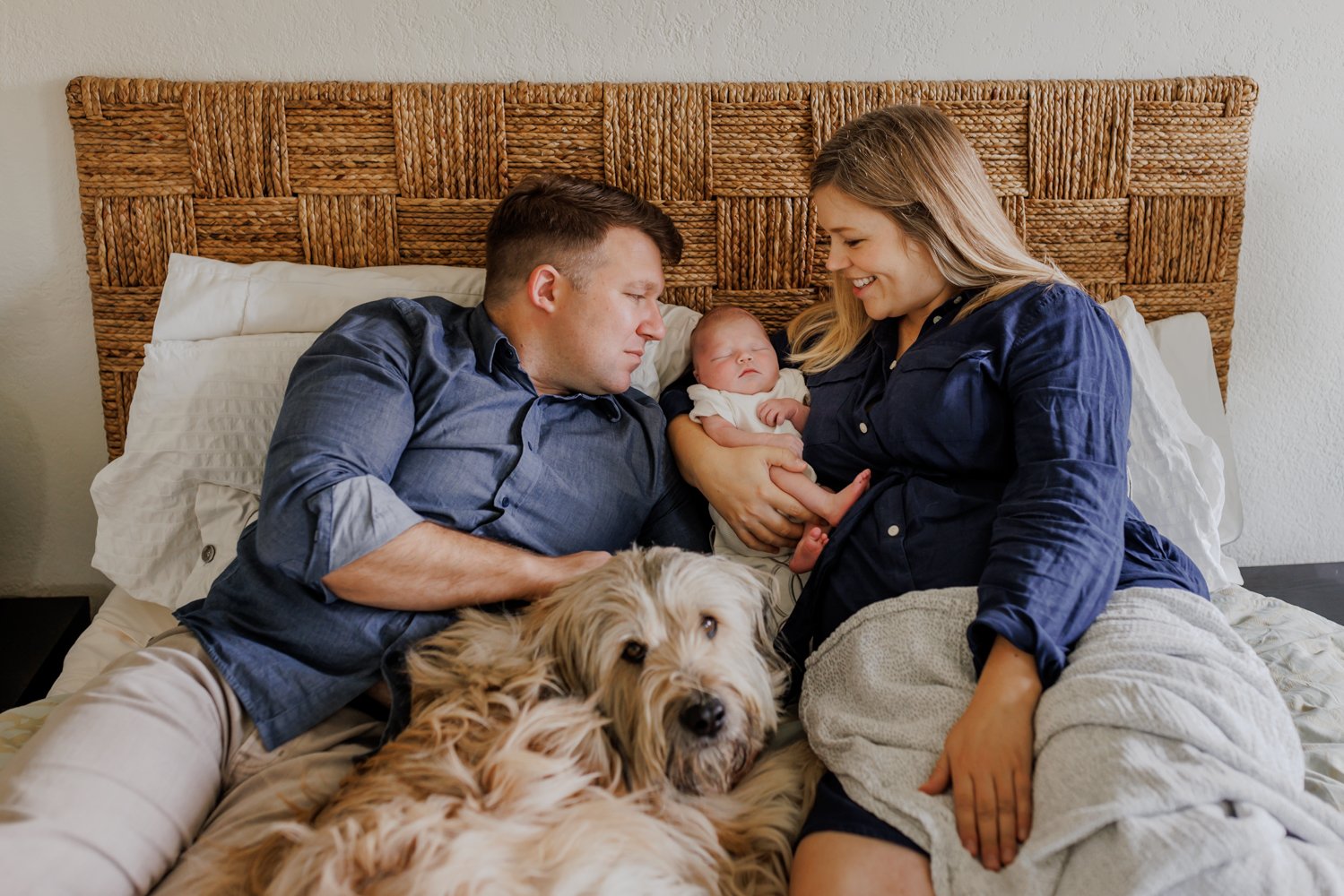 mom and dad laying on bed with their dog looking down at their newborn