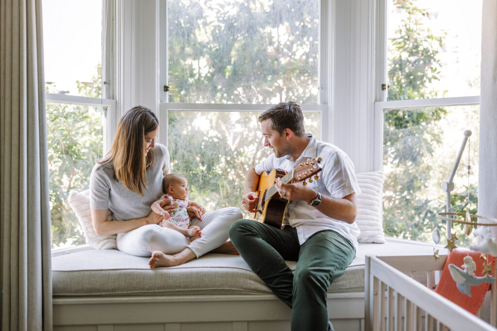 mom holding newborn at window bench seat while dad sittings next to them at home playing guitar