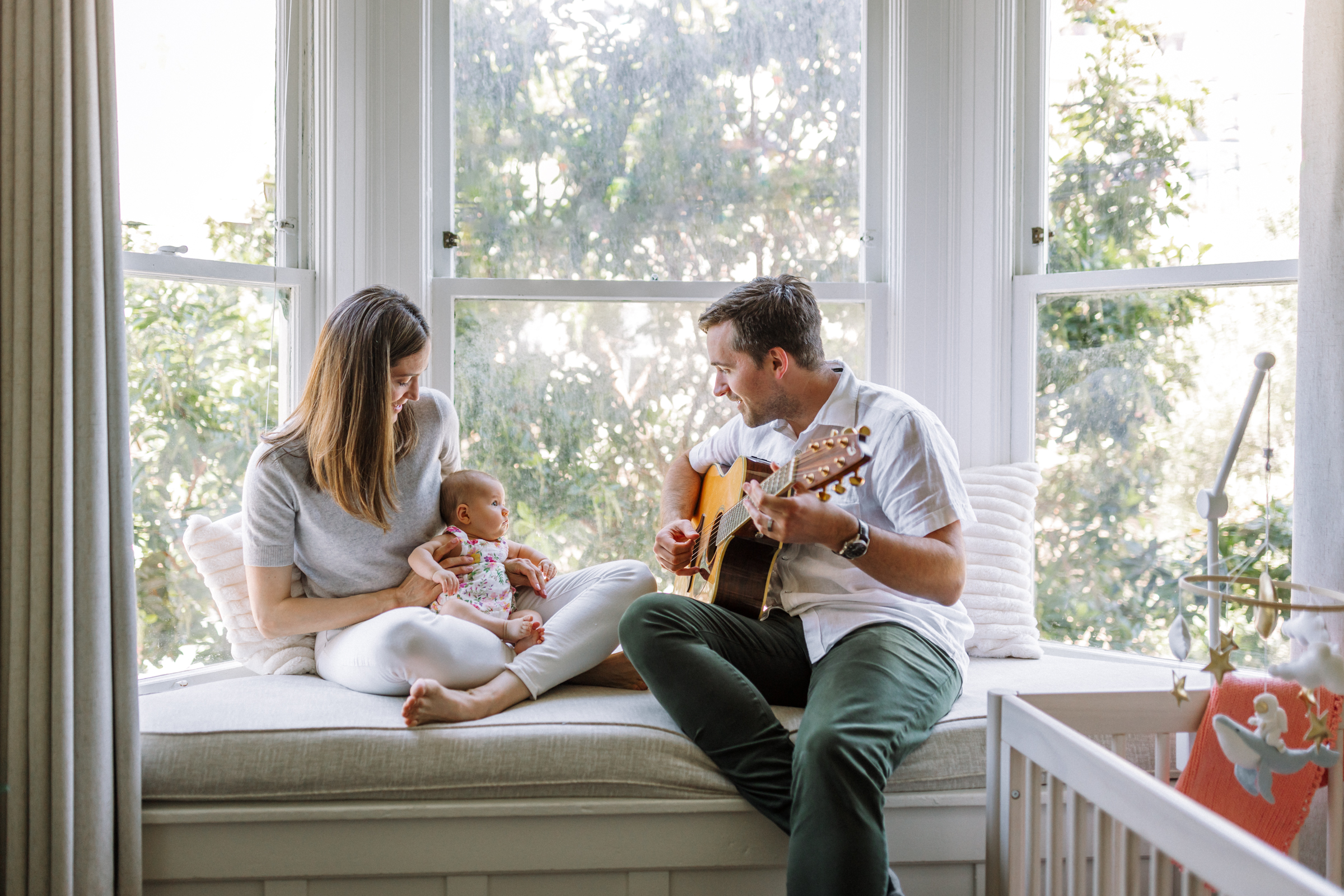 parents sitting on window bench seat with mom holding newborn as dad plays guitar