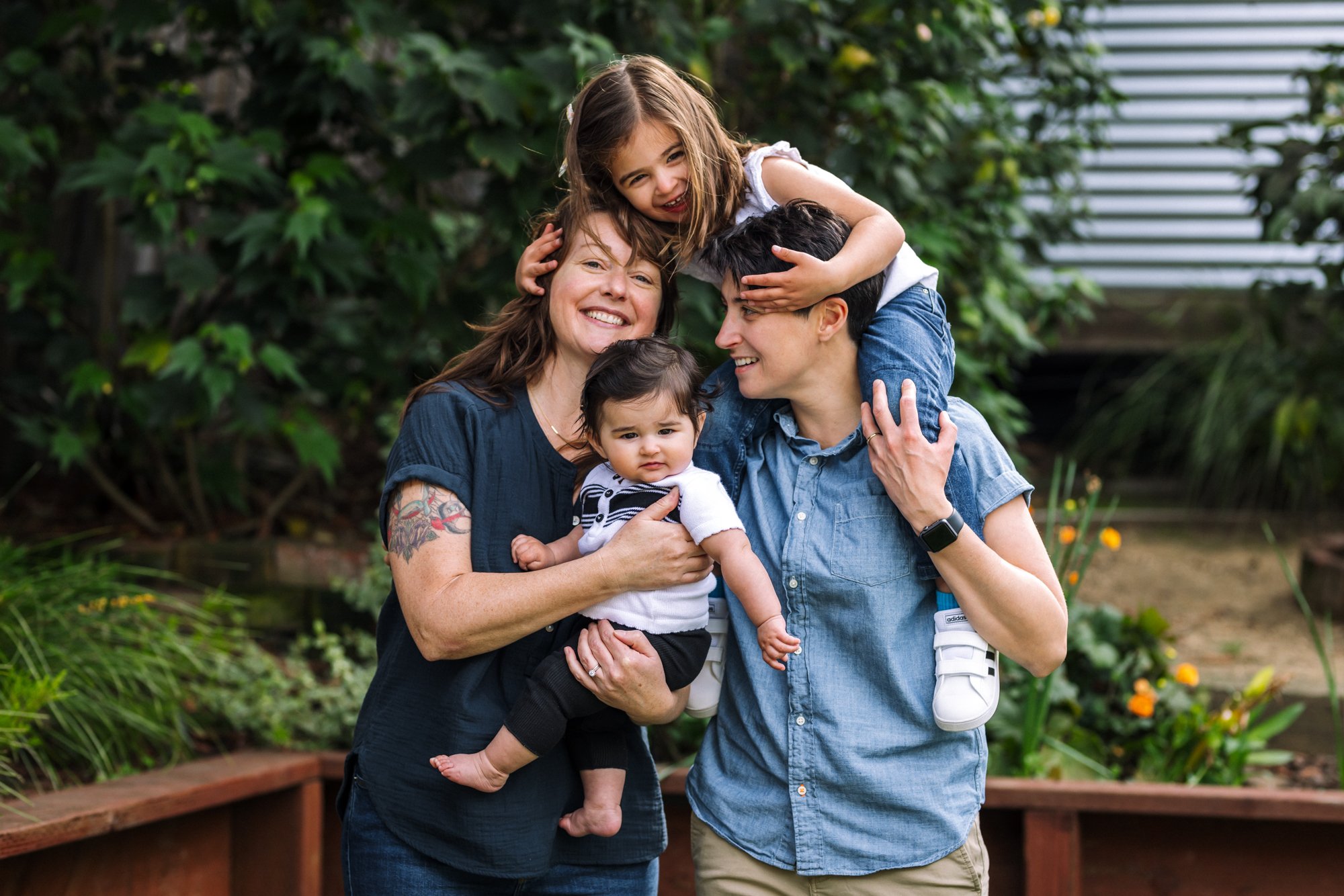 two moms holding preschool girl and baby smiling and interacting with each other at home in their backyard