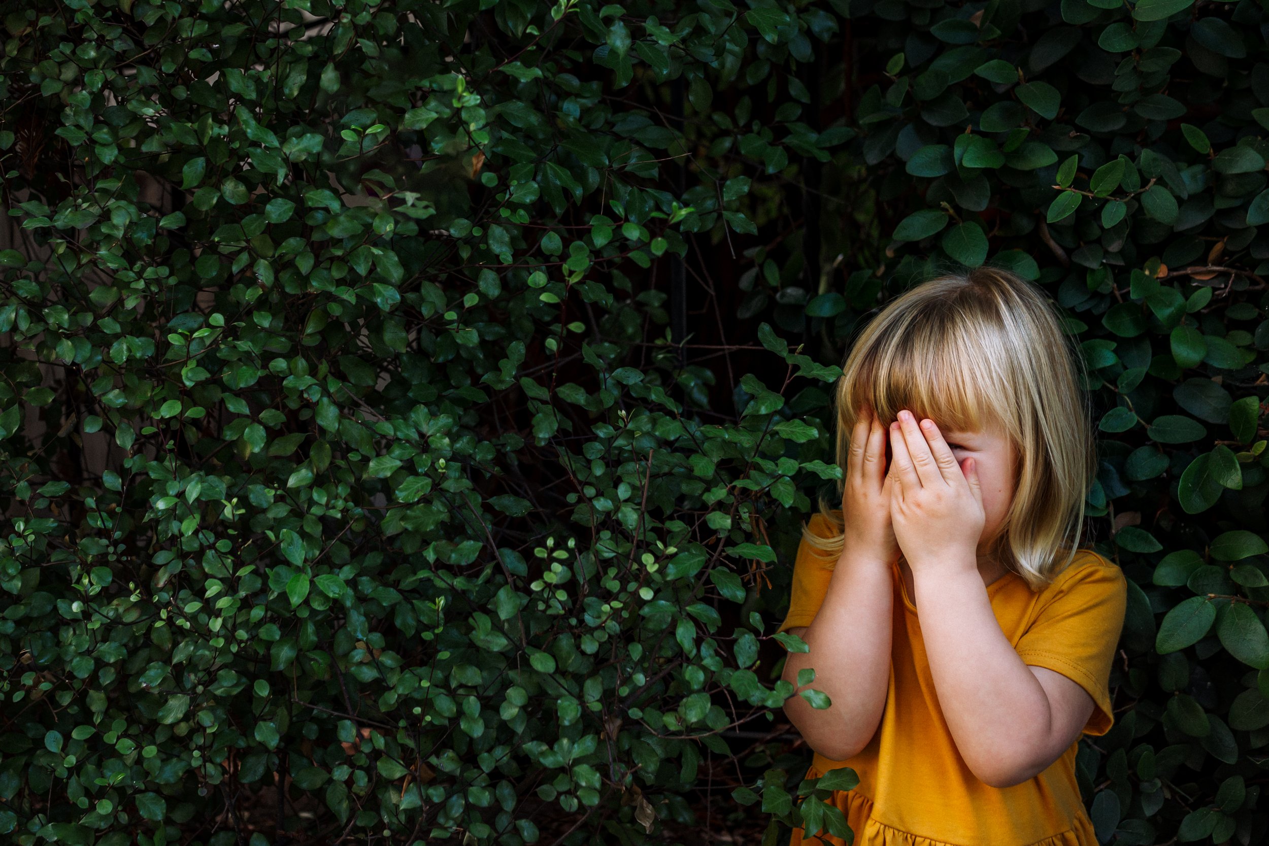 4-year-old girl in a golden dress standing in front of greenery playing peek-a-boo