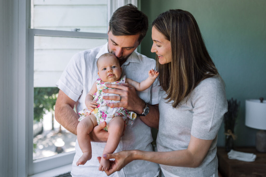 dad holding and kissing baby next to window at home while mom stands next to them smiling at newborn and holding her foot