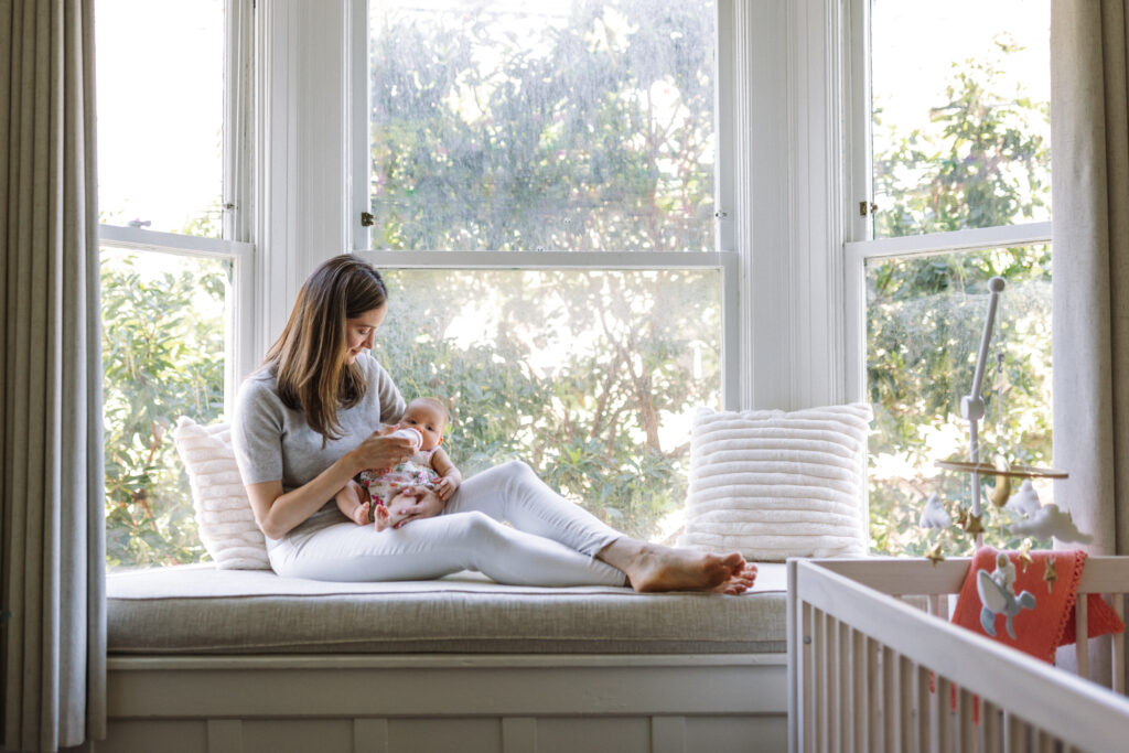 mom sitting in window bench seat holding newborn and feeing her a bottle next to the crib