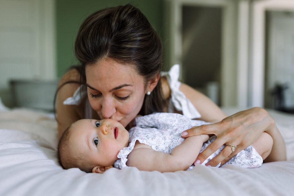 newborn laying on bed looking at camera and mom leaning over baby and kissing her cheek