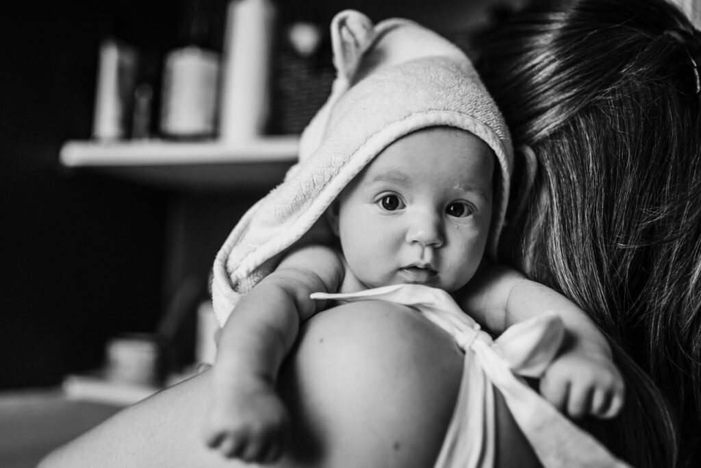 wet newborn with towel over her head draped over mom's shoulder in bathroom at home