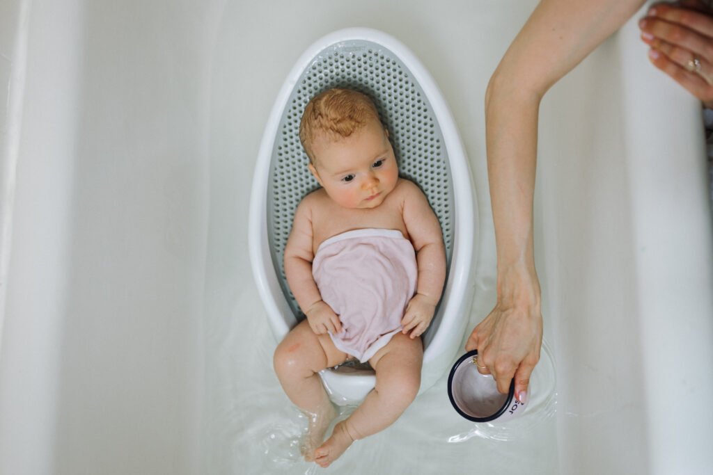 newborn in bath holder in bathtub draped in pink washcloth as mom holds a cup to pour water over baby