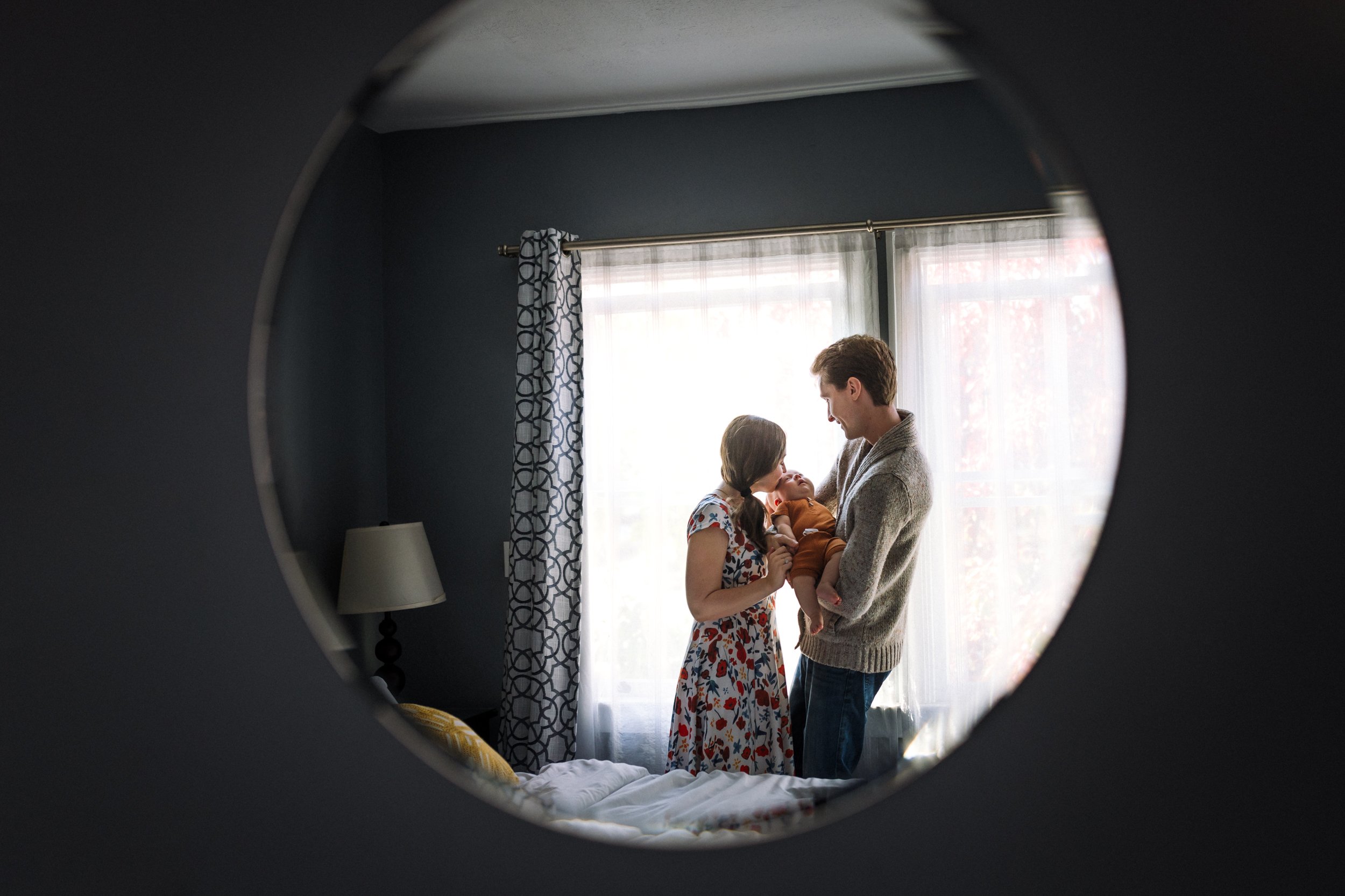 reflection image in mirror with parents standing by window with dad holding newborn and mom kissing baby