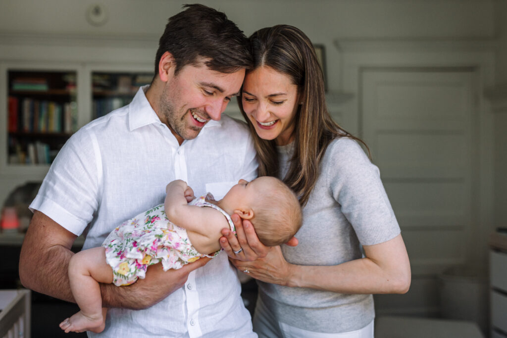 dad holding newborn with standing next to him supporting baby's head and both parents are looking down and smiling at the baby