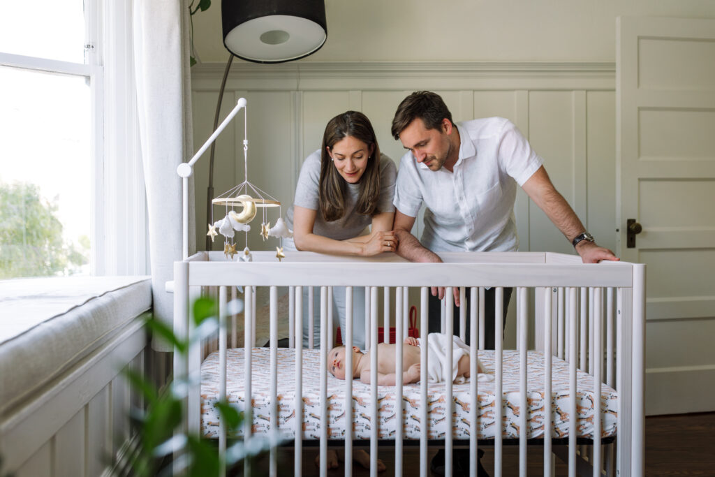 mom and dad leaning over crib at newborn with blanket draped over her as she turns to try to look towards window