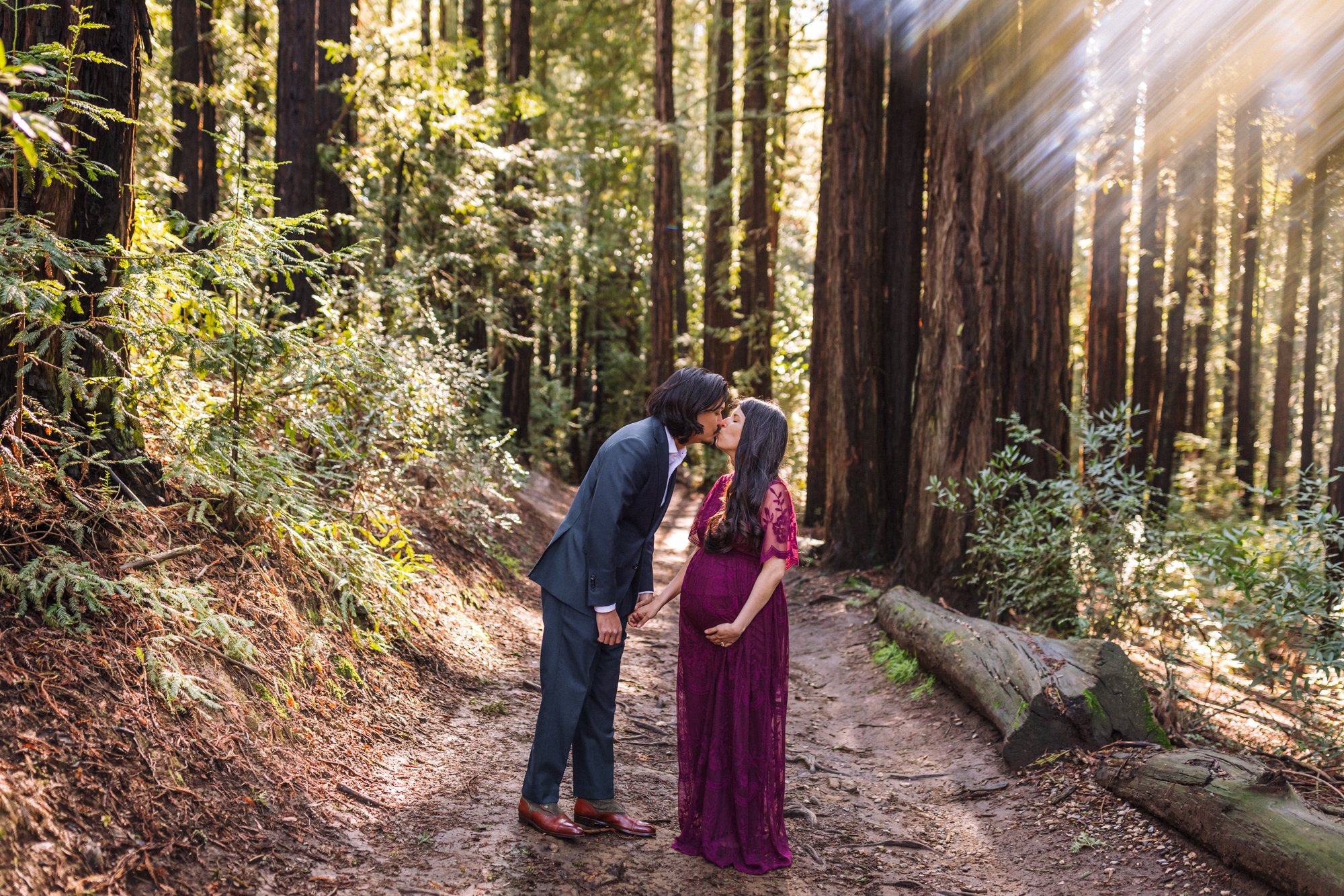 pregnant woman standing in the woods with her husband touching her baby bump as husband leans over to kiss her