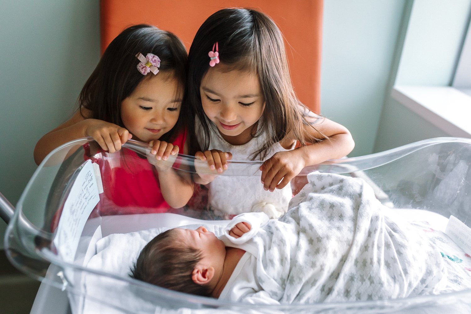 two sisters leaning over bassinet looking at newborn brother at San Francisco hospital