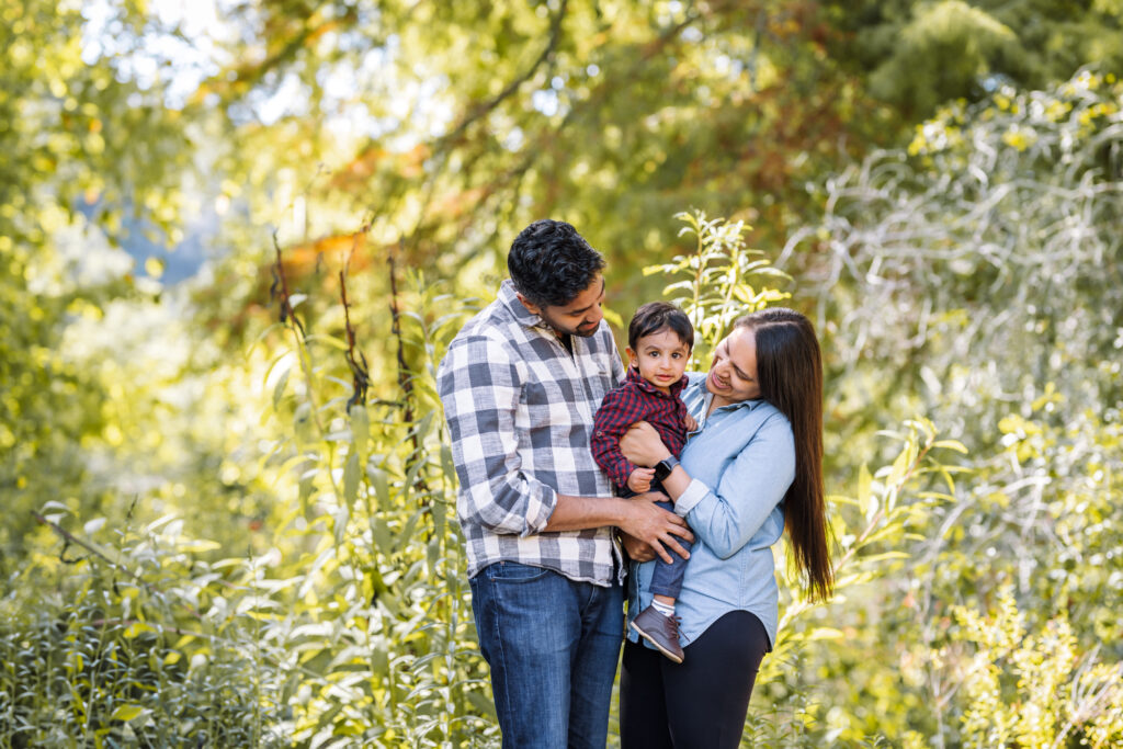 Family standing and holding son in front of greenery looking at toddler