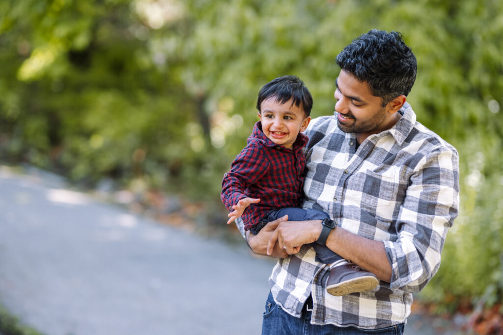 father holding toddler son and looking down at him while they both smile at a park
