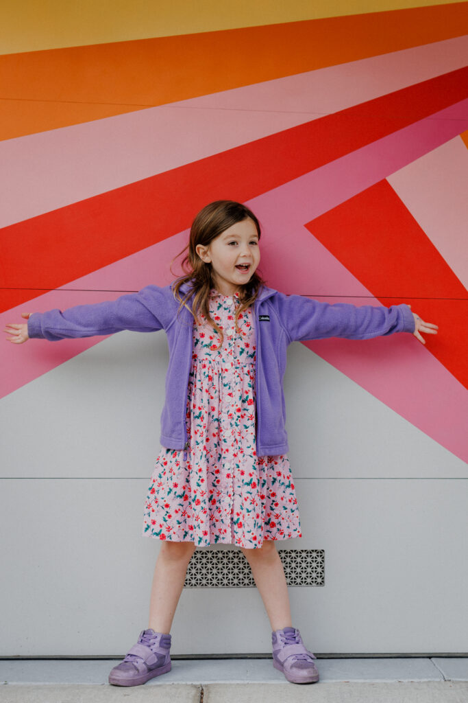Smiling girl standing with arms outstretched against a bold, colorful wall