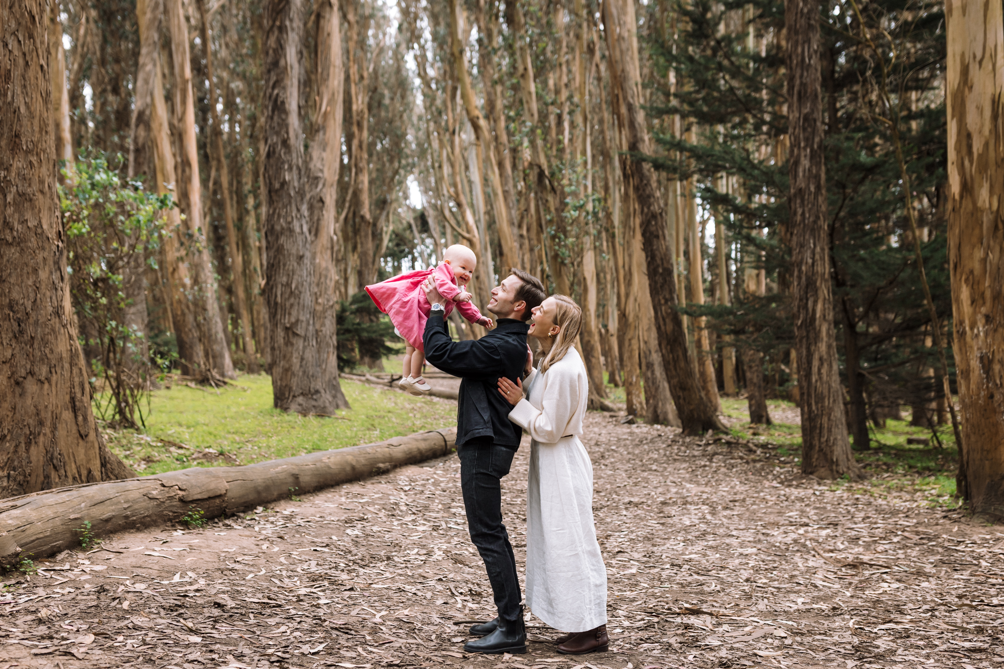 Dad holding baby up in the woods with mom looking on
