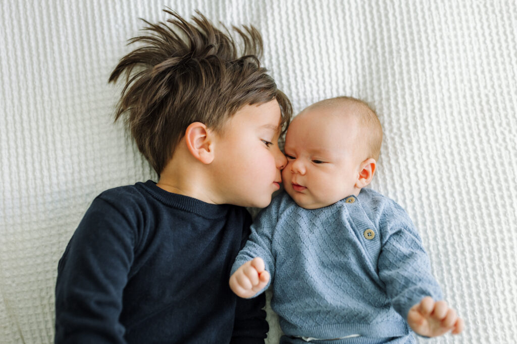 toddler kissing newborn sibling on bed in-home newborn session