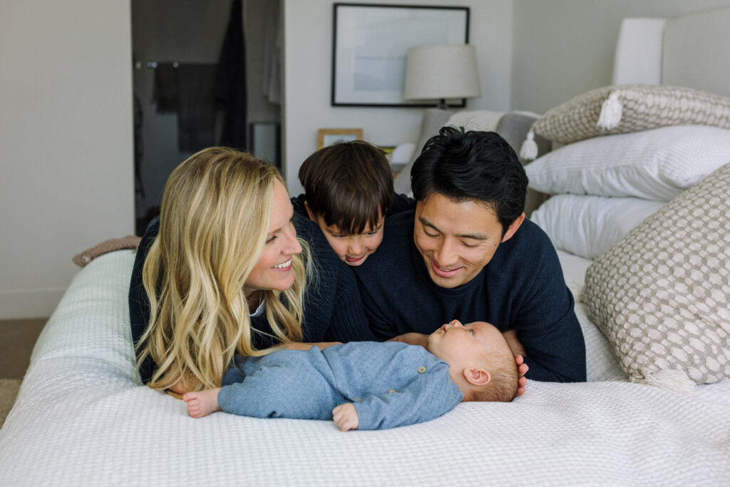 parents and toddler looking at newborn on bed in-home newborn session Alameda